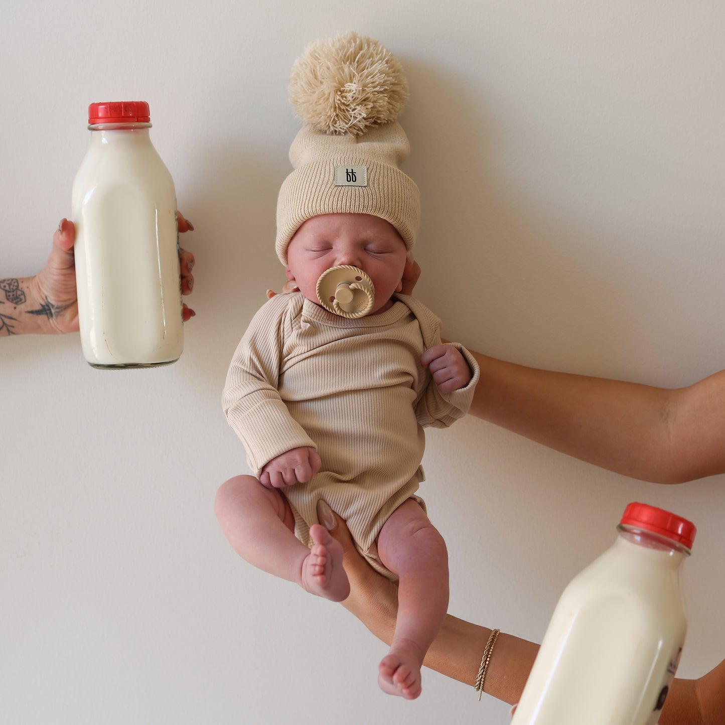 A newborn in a forever french baby ff Pom Top Knit Beanie | Oatmeal is gently cradled by adult hands, flanked by two glass milk bottles—capturing a sweet moment against a simple, light background.