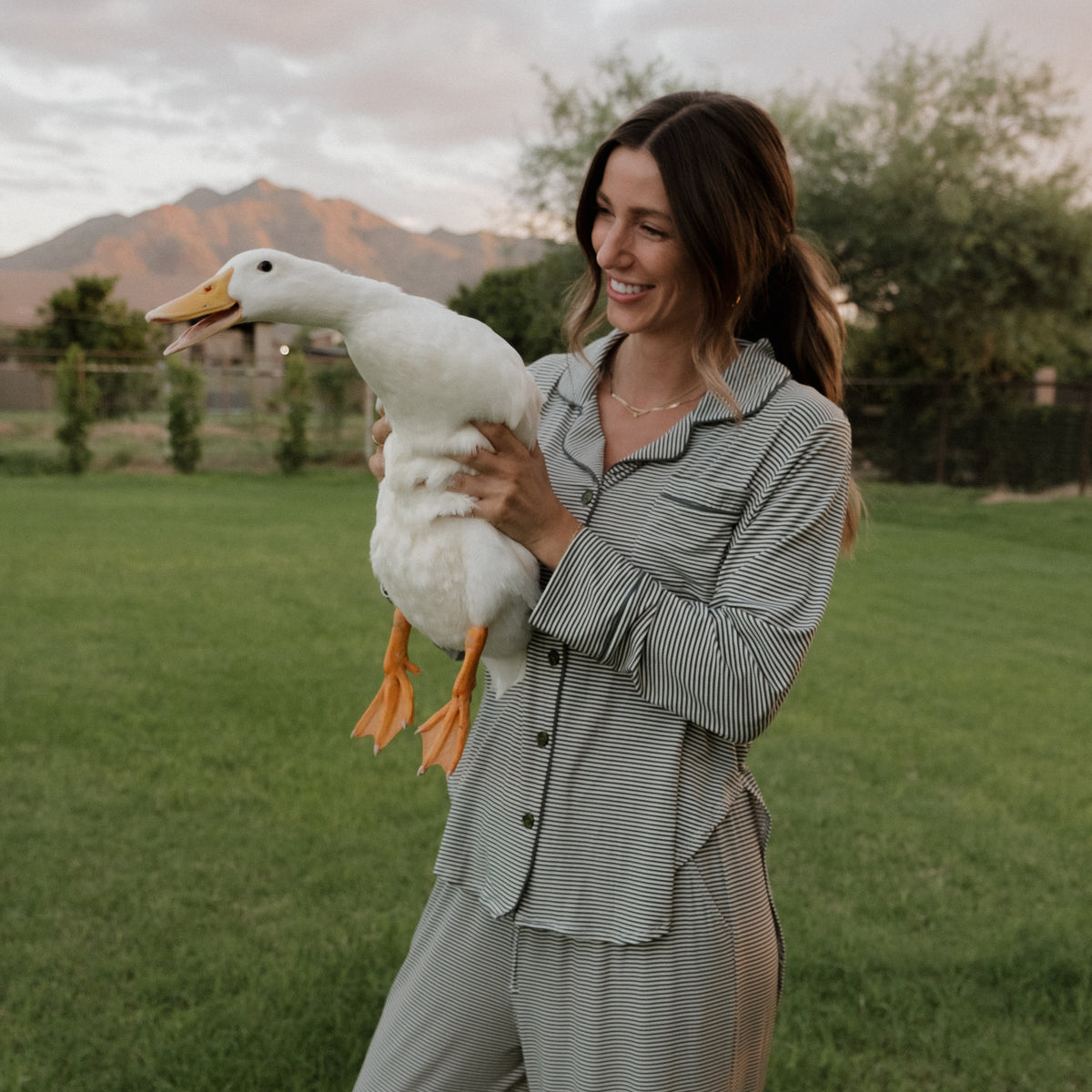 A smiling woman stands on grass holding a white duck, wearing the forever french baby Women’s Bamboo Button Up Lounge Set in Forest Stripe Ribbed, with trees and mountains in the background under a cloudy sky.