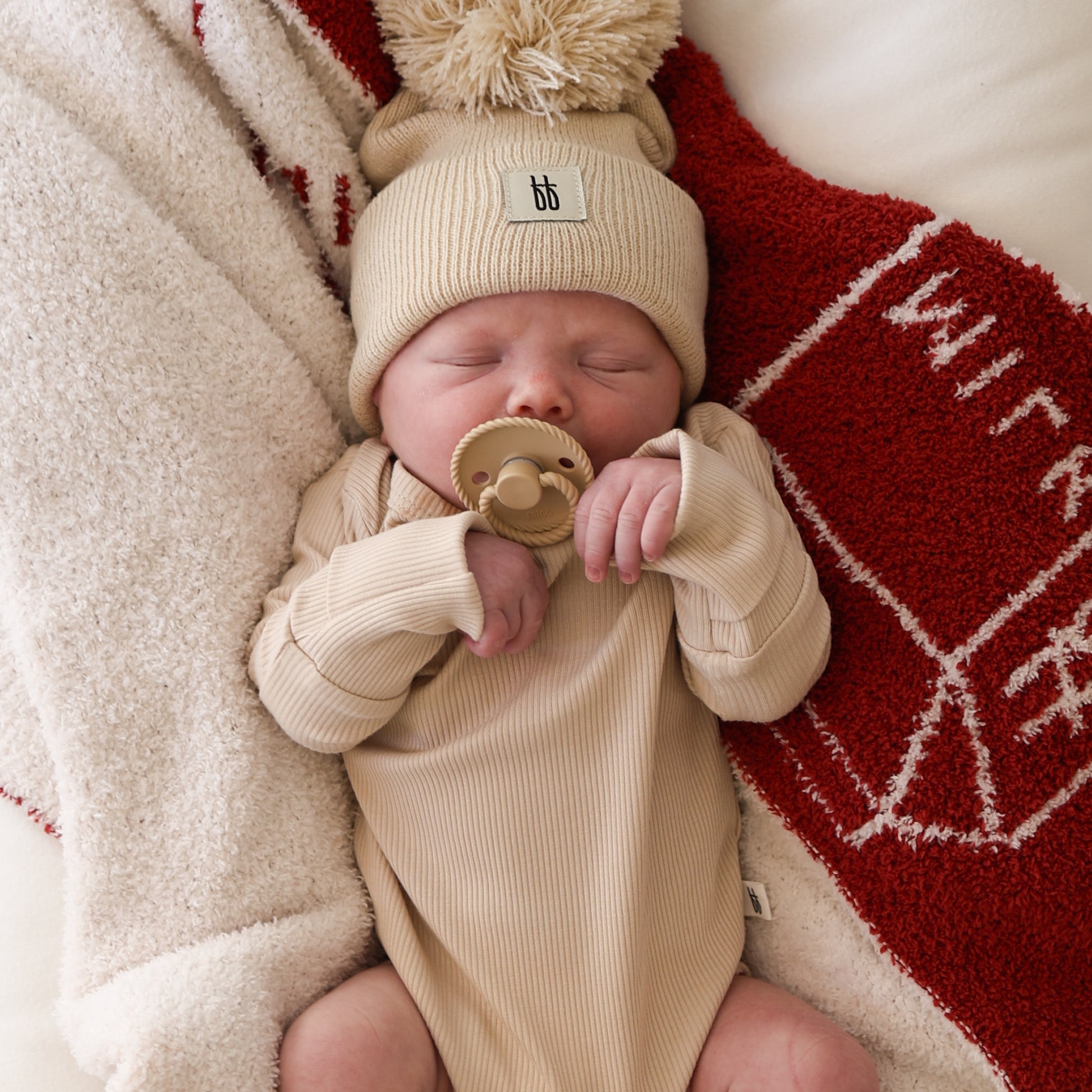 A newborn sleeps on a red and white blanket, wearing the forever french baby Long Sleeve Bamboo Snapsuit | Ribbed Cream and a matching knit hat with pom-pom, while holding a tan pacifier.