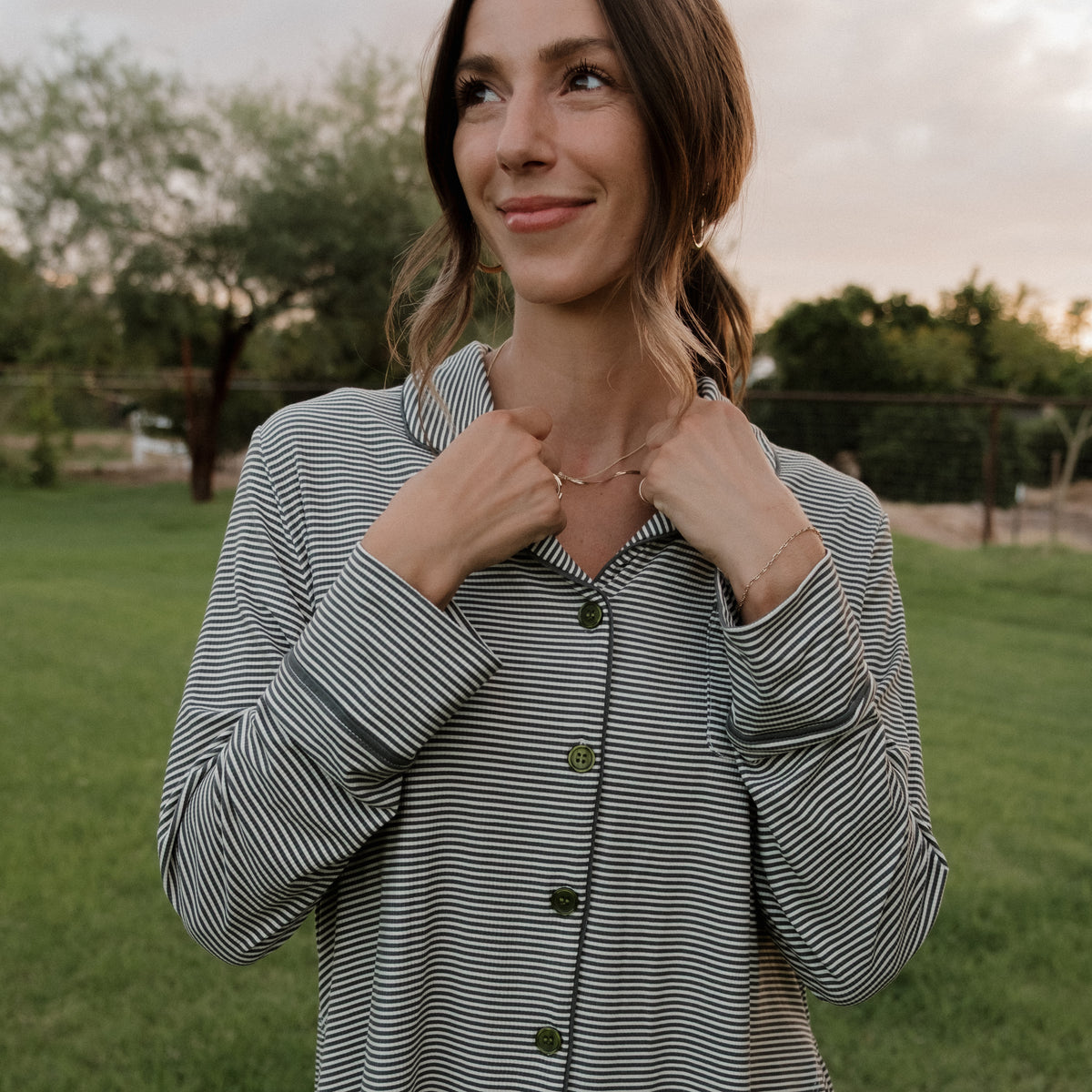 A woman stands outside on the grass at sunset, softly smiling as she gently holds a necklace. She is wearing the forever french baby Women’s Bamboo Button Up Lounge Set in Forest Stripe Ribbed, with wide-leg lounge pants and a striped button-up shirt. Trees and a fence are visible in the background.
