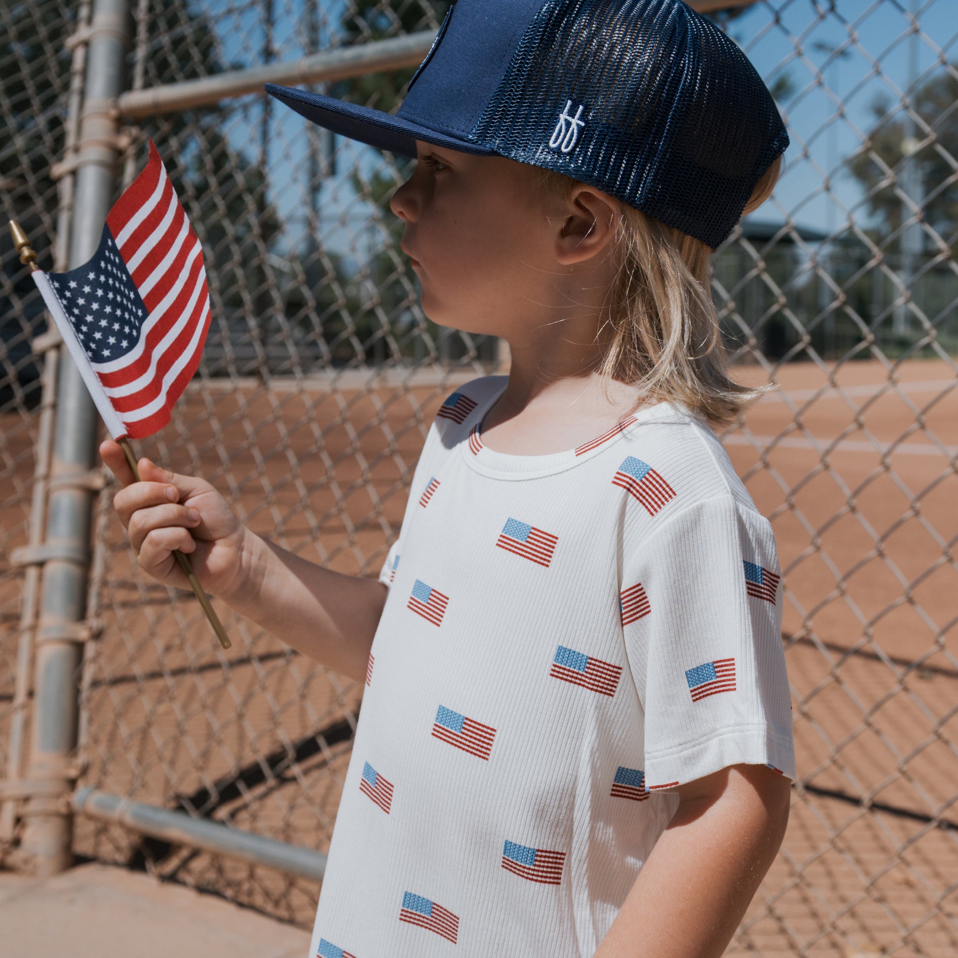A young child wears a forever french baby Bamboo Short Set | Americana Ribbed and a baseball cap, holding a small U.S. flag by a chain-link fence on a sunny day at the baseball field.
