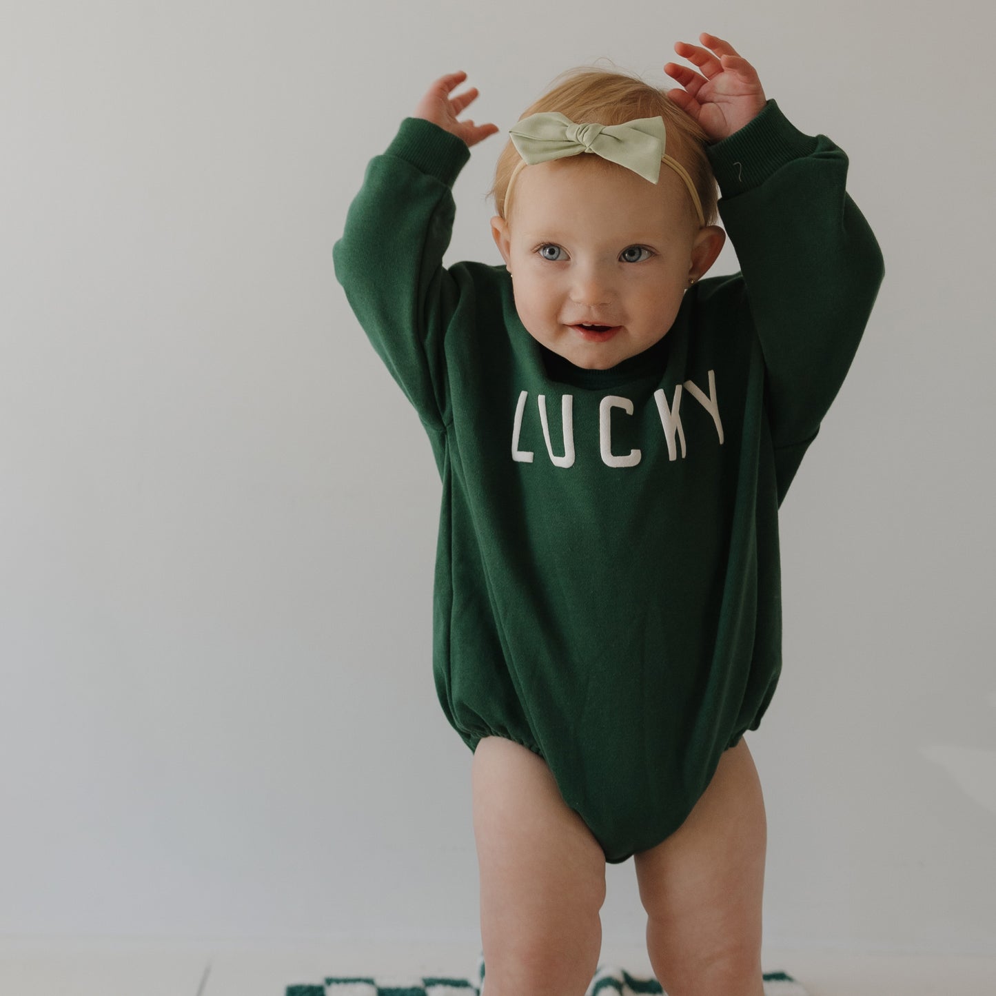 A baby wearing the forever french baby Sweatshirt Romper | Lucky and a light green bow headband raises their arms against a plain light background, dressed festively for St. Patrick’s Day.