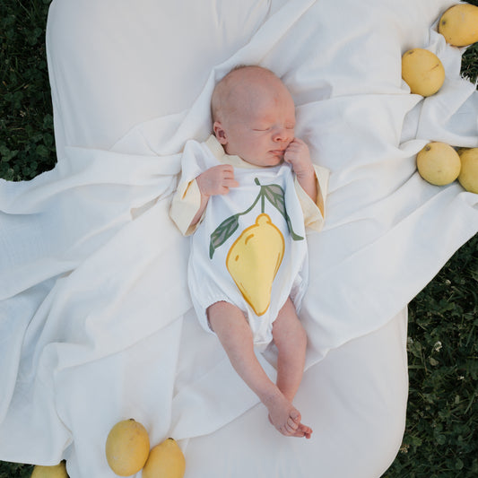 A newborn wears the forever french baby Short Sleeve Romper | Lemon, featuring a bold yellow lemon print, lying on a white blanket with fresh lemons on green grass—a perfect summer outfit.