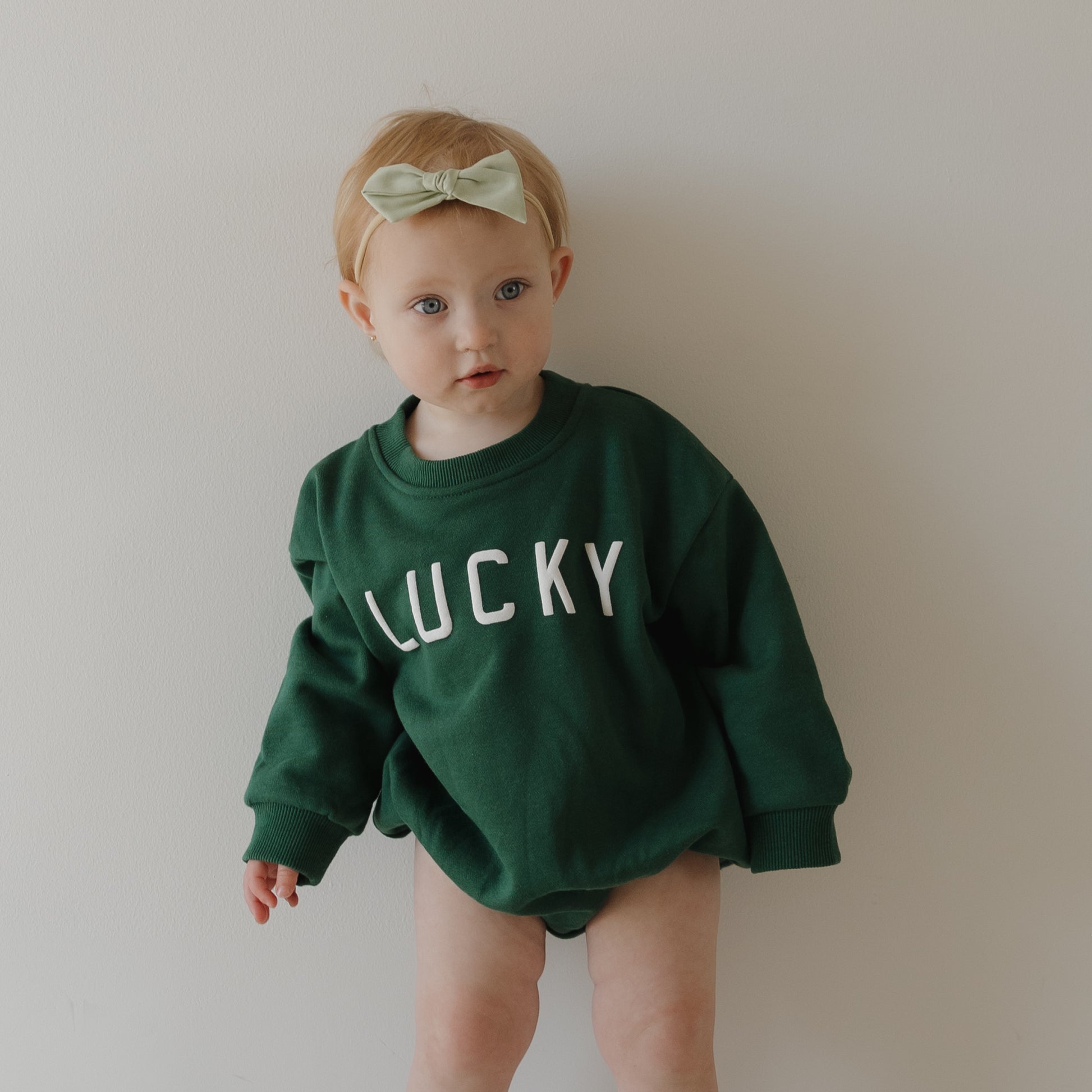 A young child wears the forever french baby Sweatshirt Romper | Lucky and a bow headband, standing by a plain wall—an adorable pick for St. Patrick’s Day.