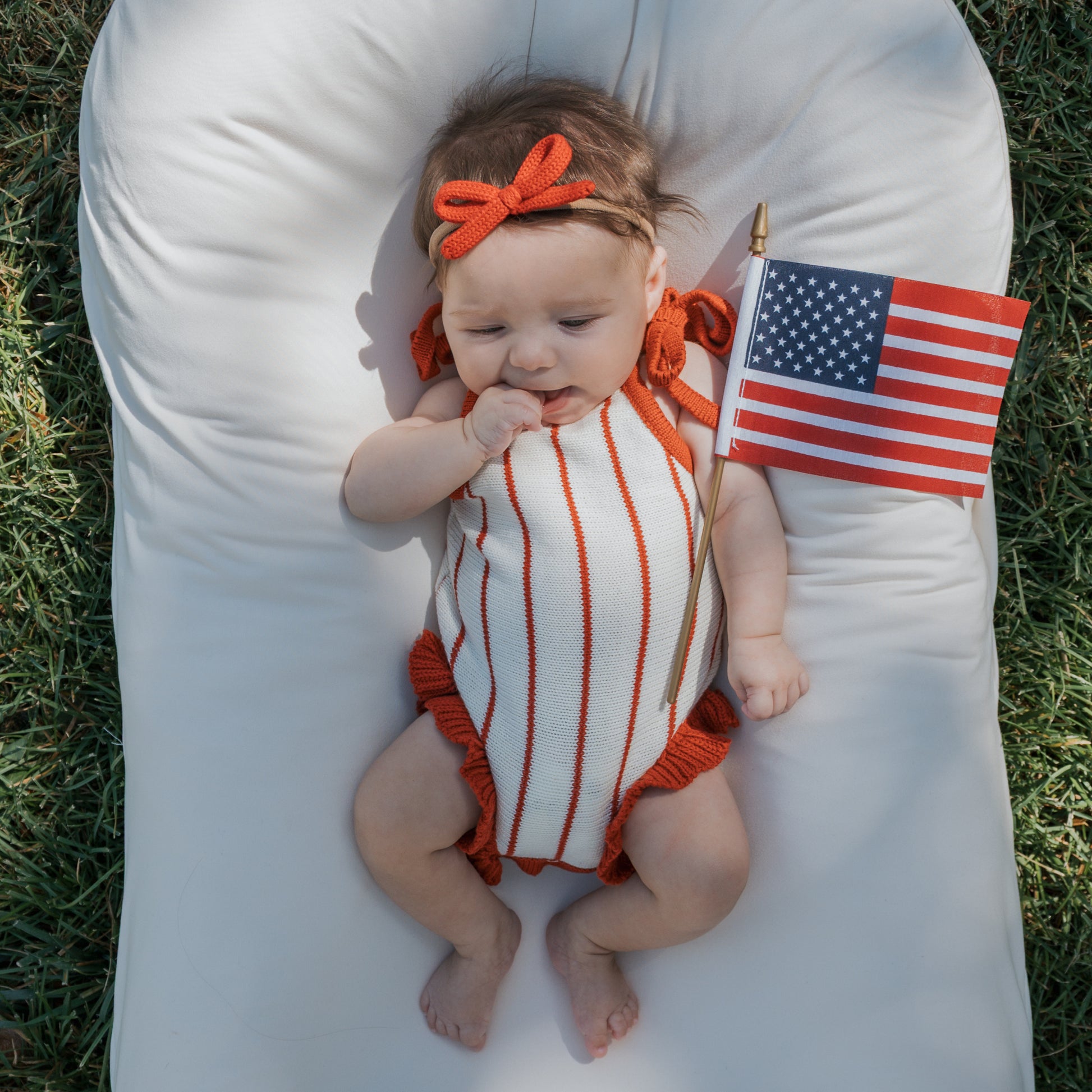 A baby in the forever french baby Knit Ruffle Romper | Picnic Pinstripe and a red headband lies on a white cushion outdoors, holding a small American flag, with grass visible beneath.