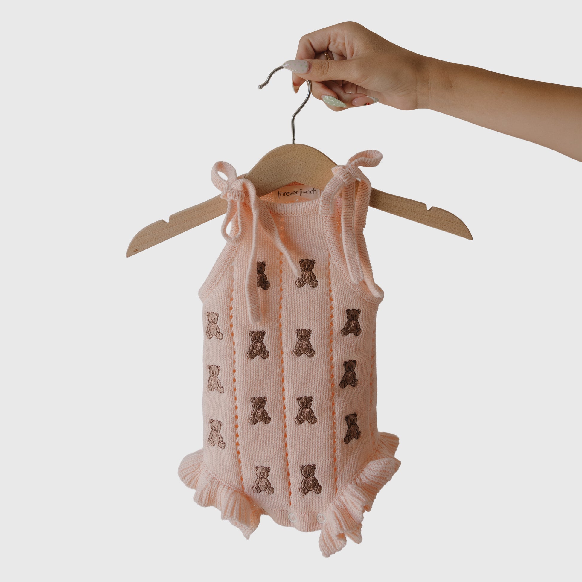 A hand holds the forever french baby Knit Ruffle Romper | Lottie, a pink cotton romper with brown teddy bear patterns and ruffled leg openings, displayed on a wooden hanger against a plain white background.