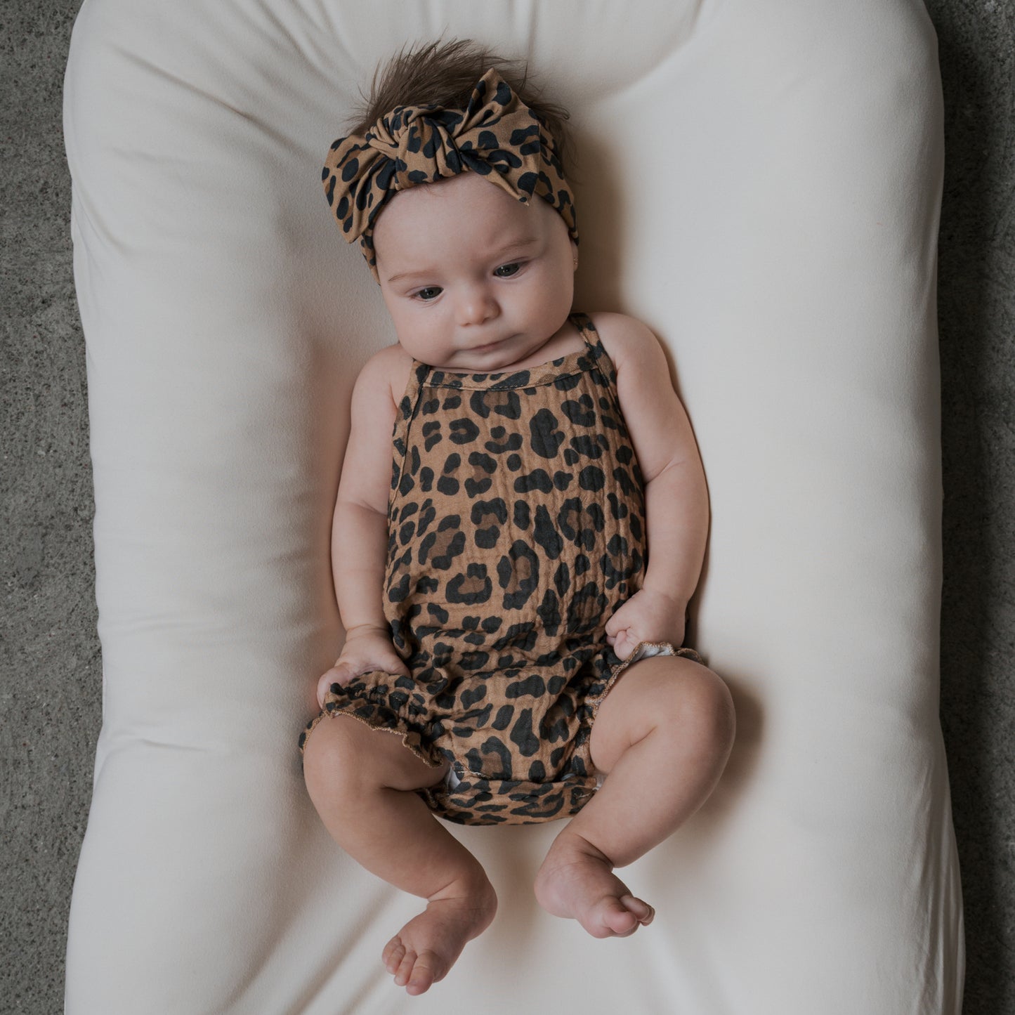 A baby lies on a white cushioned surface wearing the forever french baby Muslin Sleeveless Tank Romper in Leopard print with a matching headband, looking down thoughtfully.