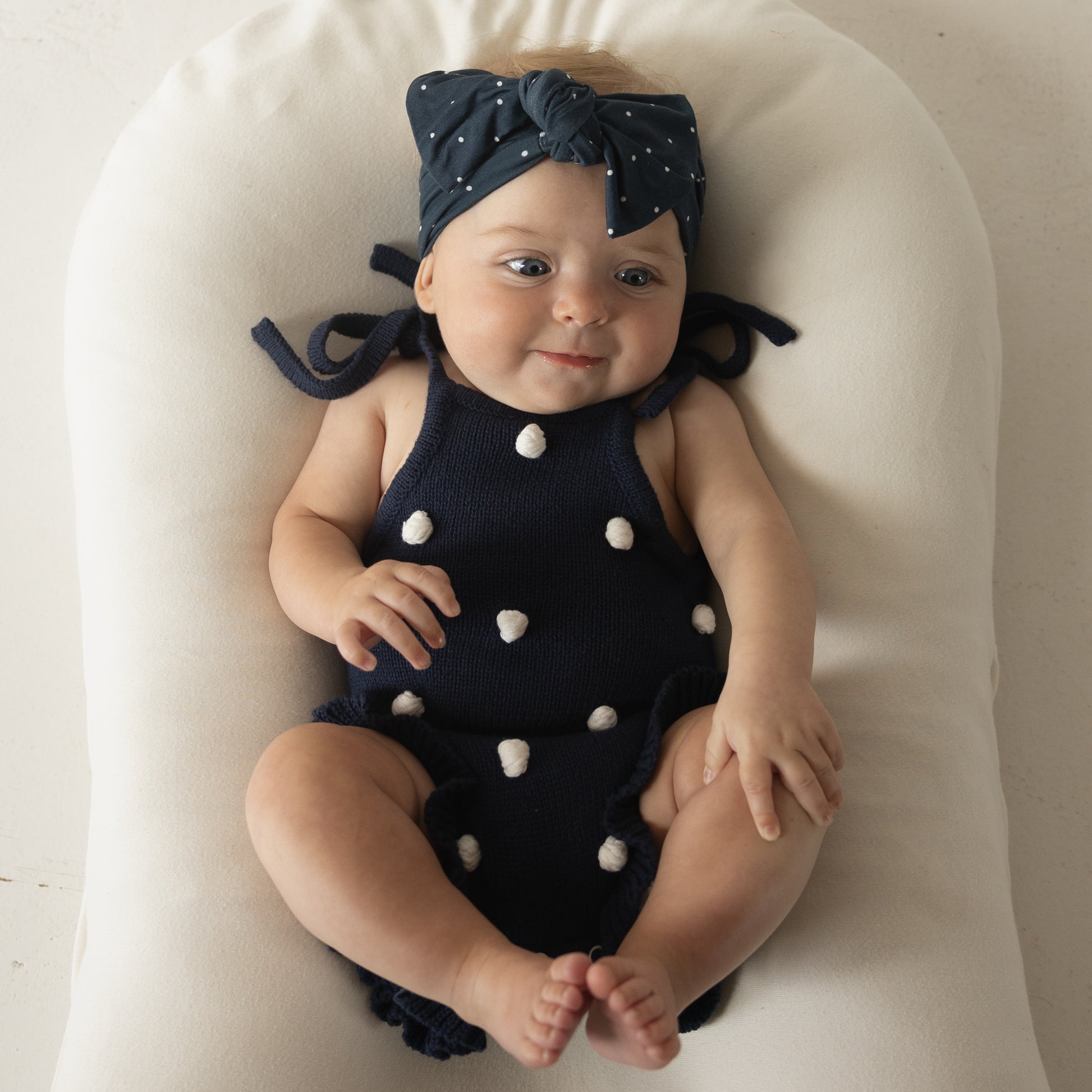 A baby in the forever french baby Knit Ruffle Romper | Emery, featuring soft knit fabric and convenient snap closures, paired with a matching navy blue polka dot headband, lies smiling on a white cushioned surface.