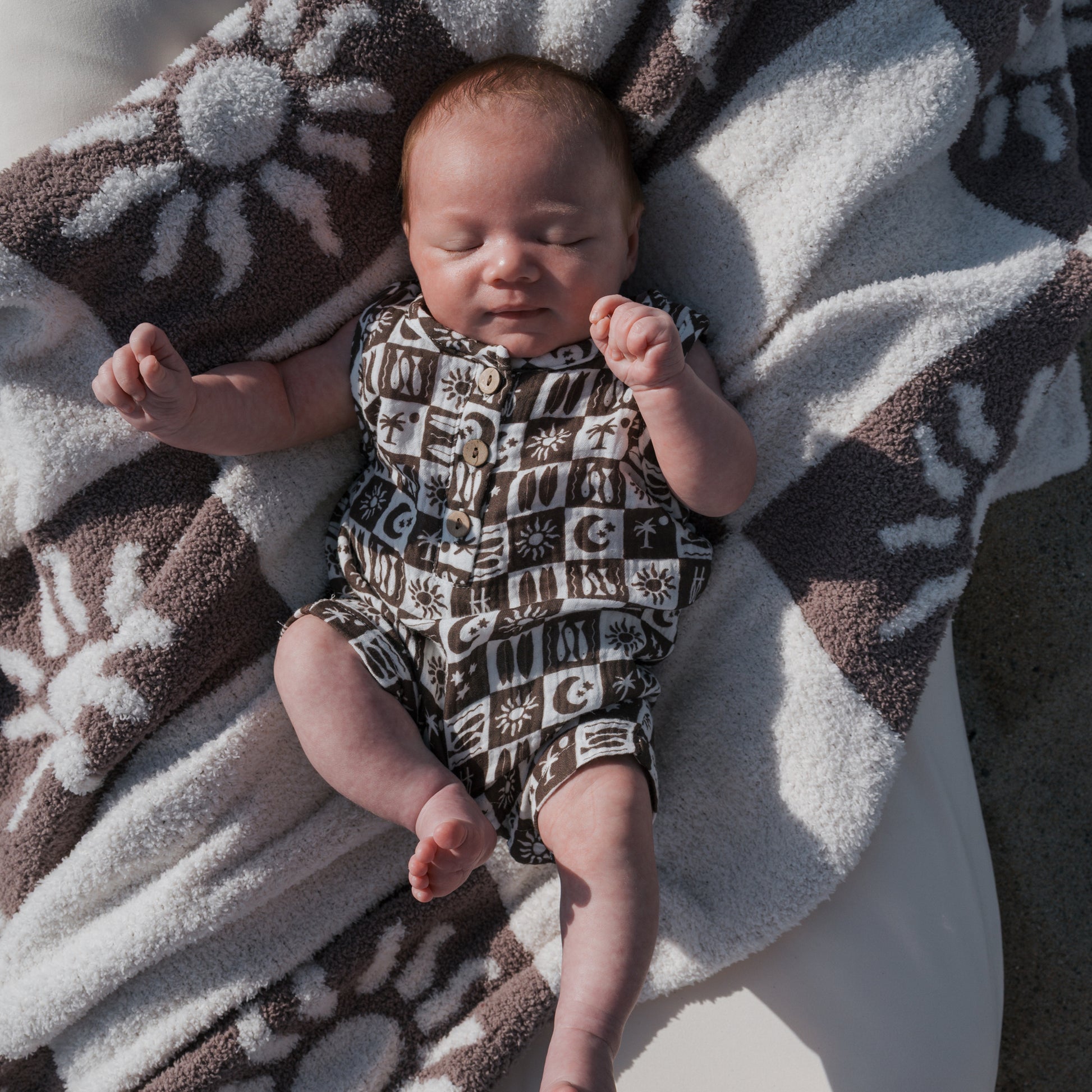 A baby in a forever french baby Muslin Tank Romper | Summer Check lies on a sun and moon-themed blanket outdoors, eyes closed and arms raised, relaxing in natural light.
