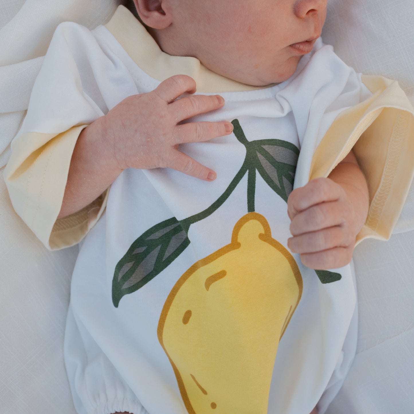 A baby in the forever french baby Short Sleeve Romper | Lemon, featuring soft cotton with beige trim and a bold lemon graphic, rests on a white blanket, one hand on their chest.
