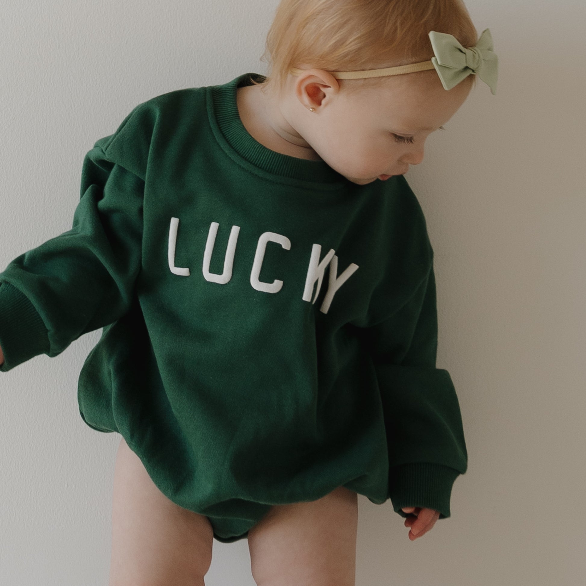 A baby with light hair and a green bow wears the forever french baby "Sweatshirt Romper | Lucky." She looks down, ready for St. Patrick’s Day, against a plain, light background.