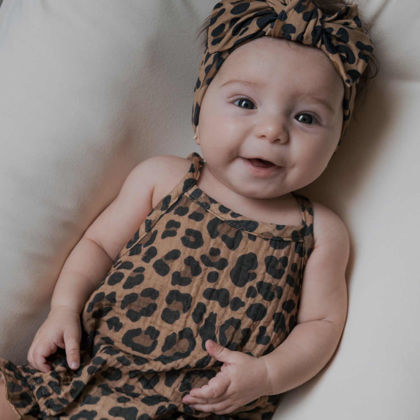 A smiling baby lies on a light surface, wearing the Muslin Sleeveless Tank Romper in Leopard from forever french baby, paired with a matching bow headband. The newborn cotton outfit highlights the baby's bright eyes and open mouth.