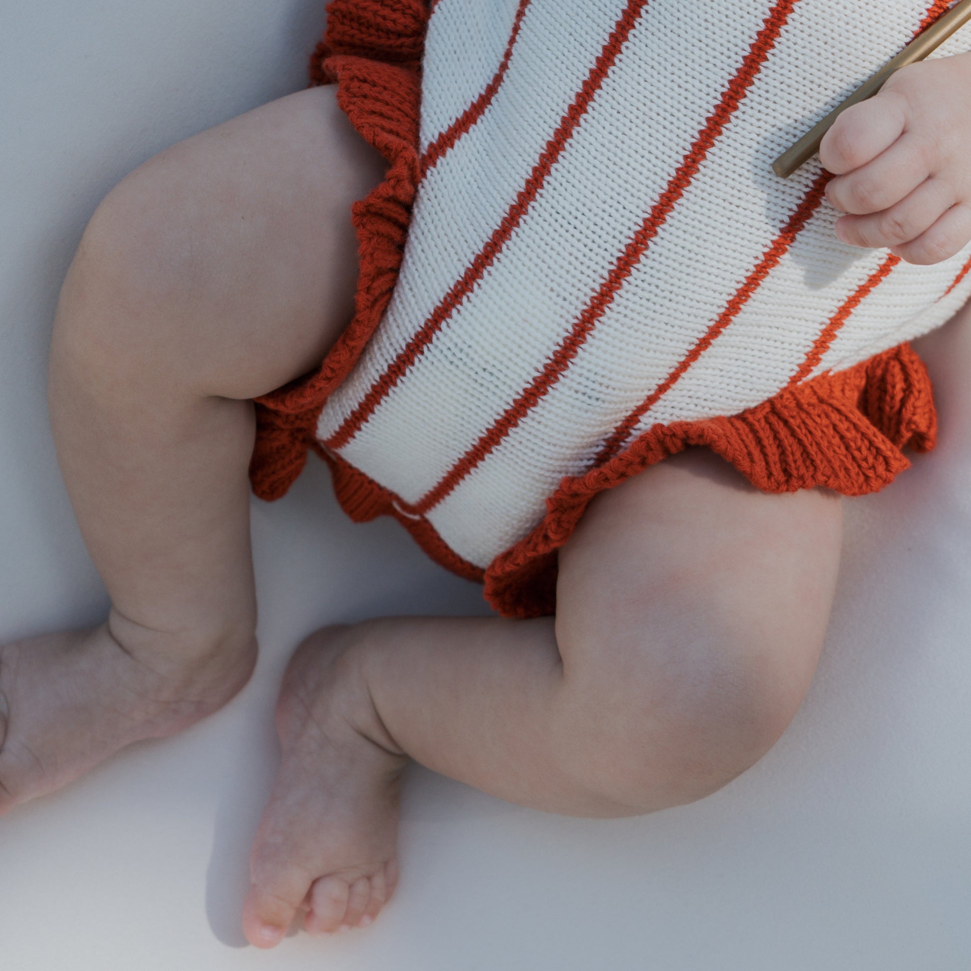 A baby in the forever french baby Knit Ruffle Romper | Picnic Pinstripe is lying down, showing only legs, feet, and a hand. The cozy romper features red and white stripes and is made from 100% cotton.
