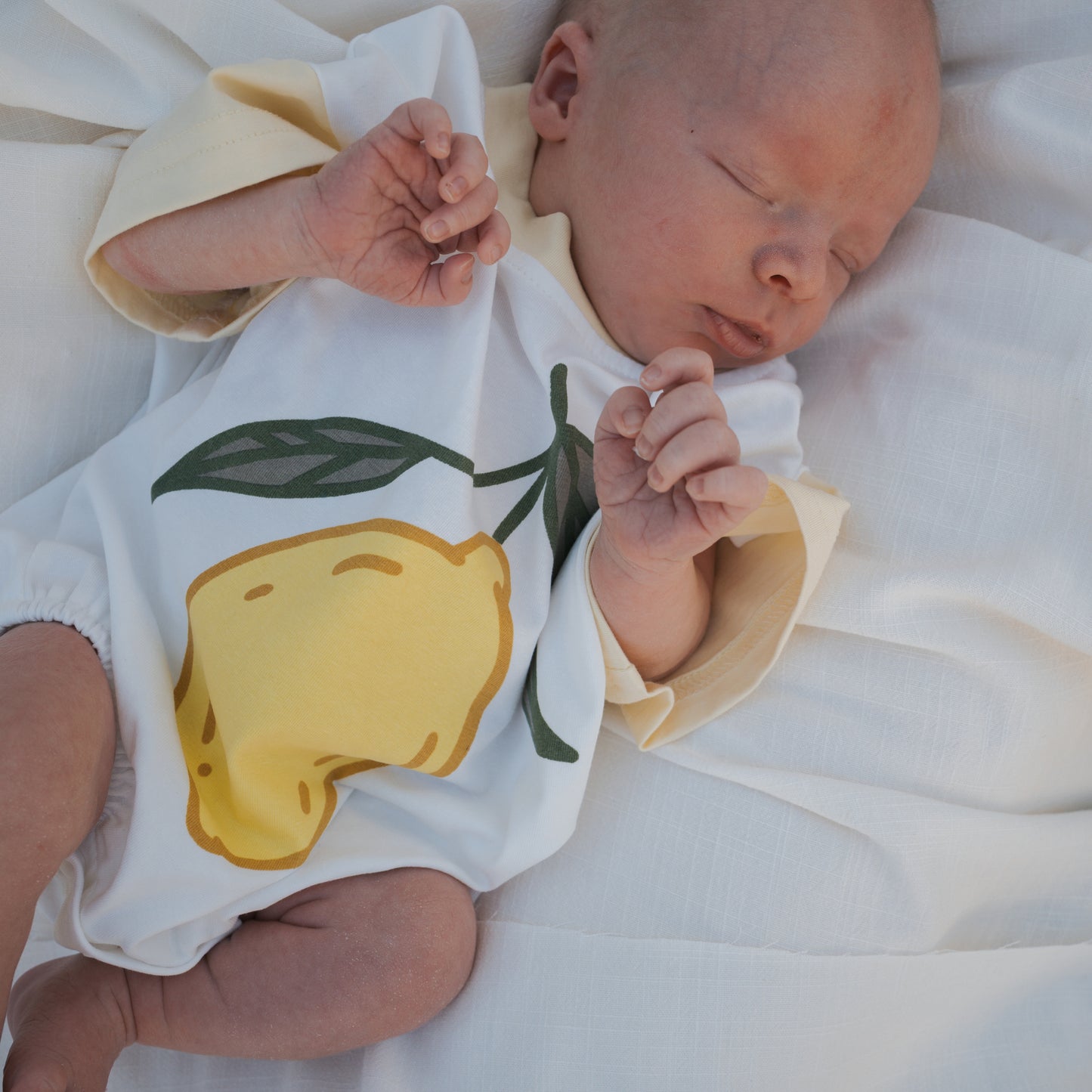 A newborn sleeps on a white blanket, wearing the forever french baby Short Sleeve Romper | Lemon, featuring a soft cotton fabric with a large yellow lemon and green leaves printed on the front.