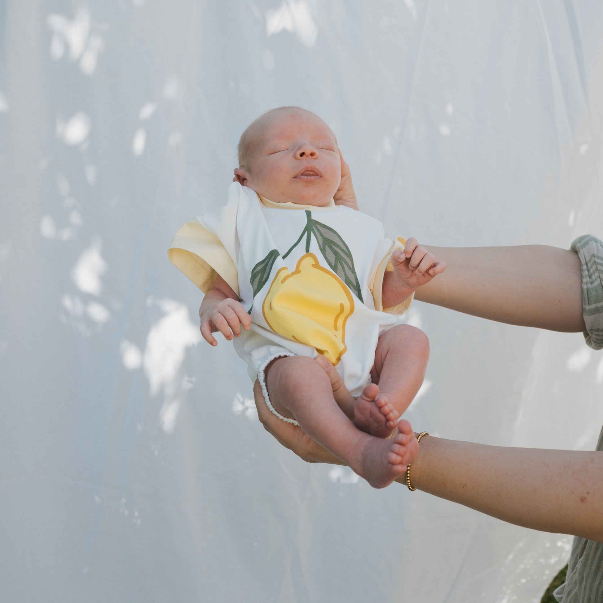 A newborn in the forever french baby Short Sleeve Romper | Lemon is cradled outdoors amid dappled sunlight—a perfect summer outfit for those first precious days.