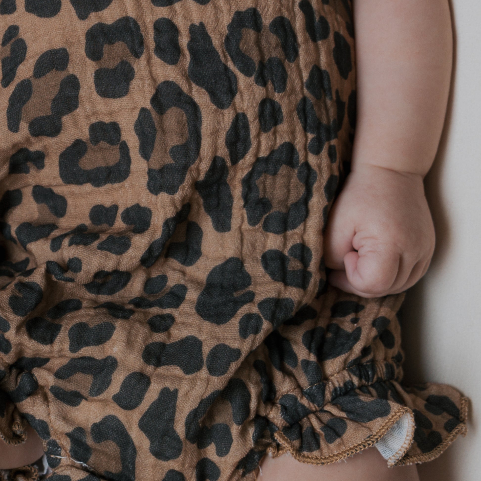 A close-up of a baby wearing the Muslin Sleeveless Tank Romper | Leopard by forever french baby, showing a black leopard print on soft cotton, with the baby's arm and clenched fist resting by their side.