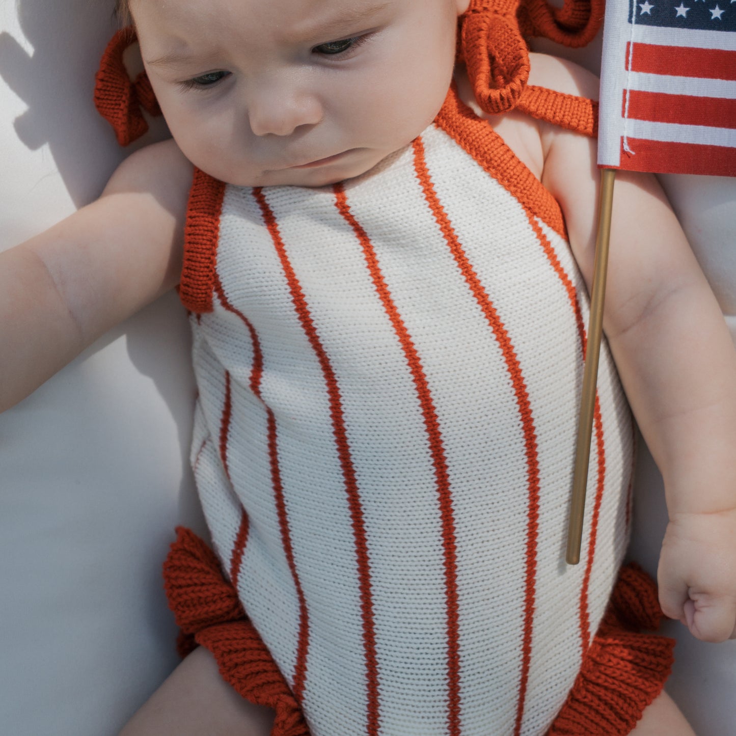 A baby in the forever french baby Knit Ruffle Romper | Picnic Pinstripe—a white romper with red stripes and trim—holds a small American flag while lying down, gazing down thoughtfully.