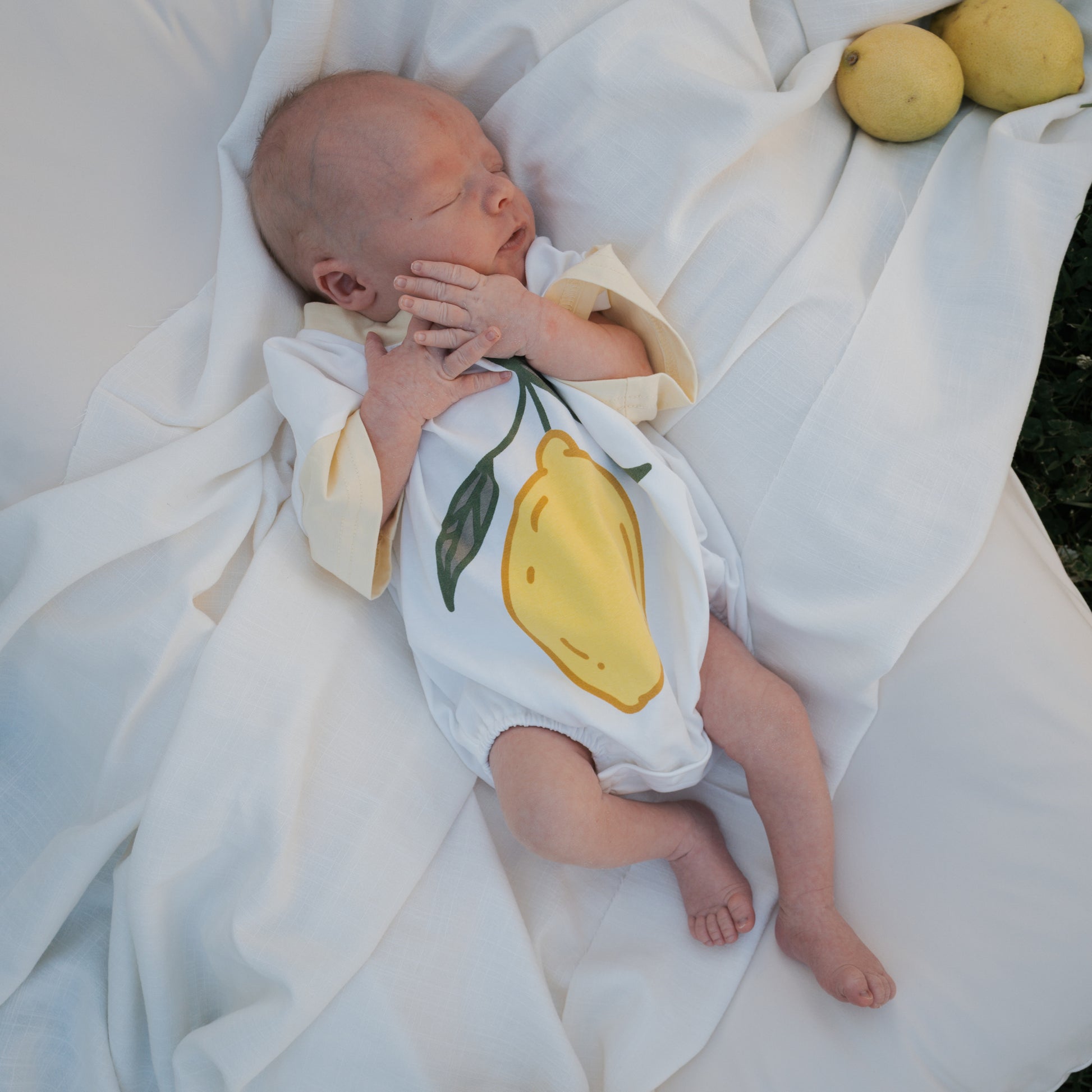 A newborn in the forever french baby Short Sleeve Romper | Lemon lies on a white blanket beside two lemons, showing off this playful yellow print—perfect for a summer baby outfit.