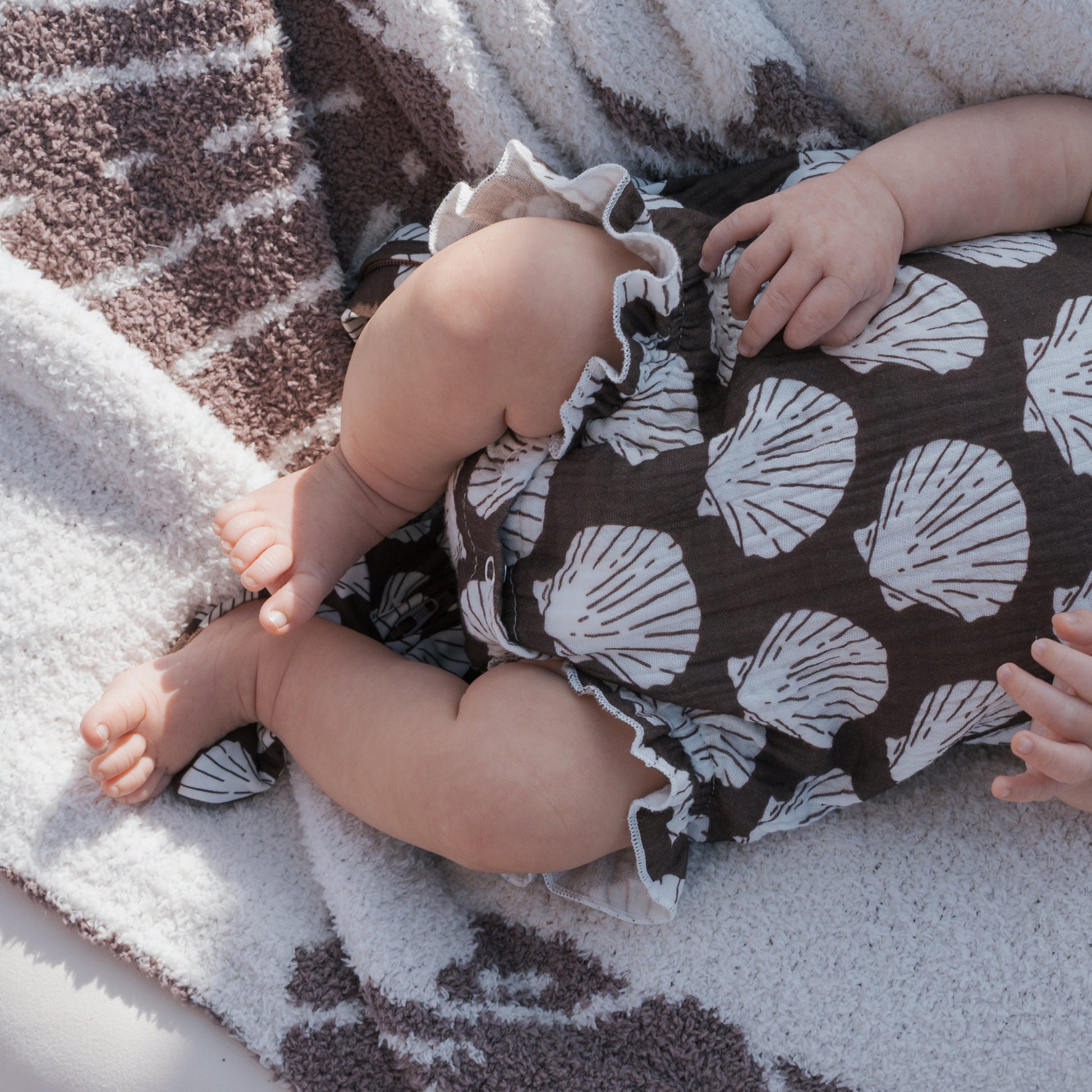 A baby wears the forever french baby Muslin Sleeveless Tank Romper | Seashells, featuring a brown color with white shell patterns, while lying on a soft blanket. The baby's chubby legs and hands are visible; face is not shown.