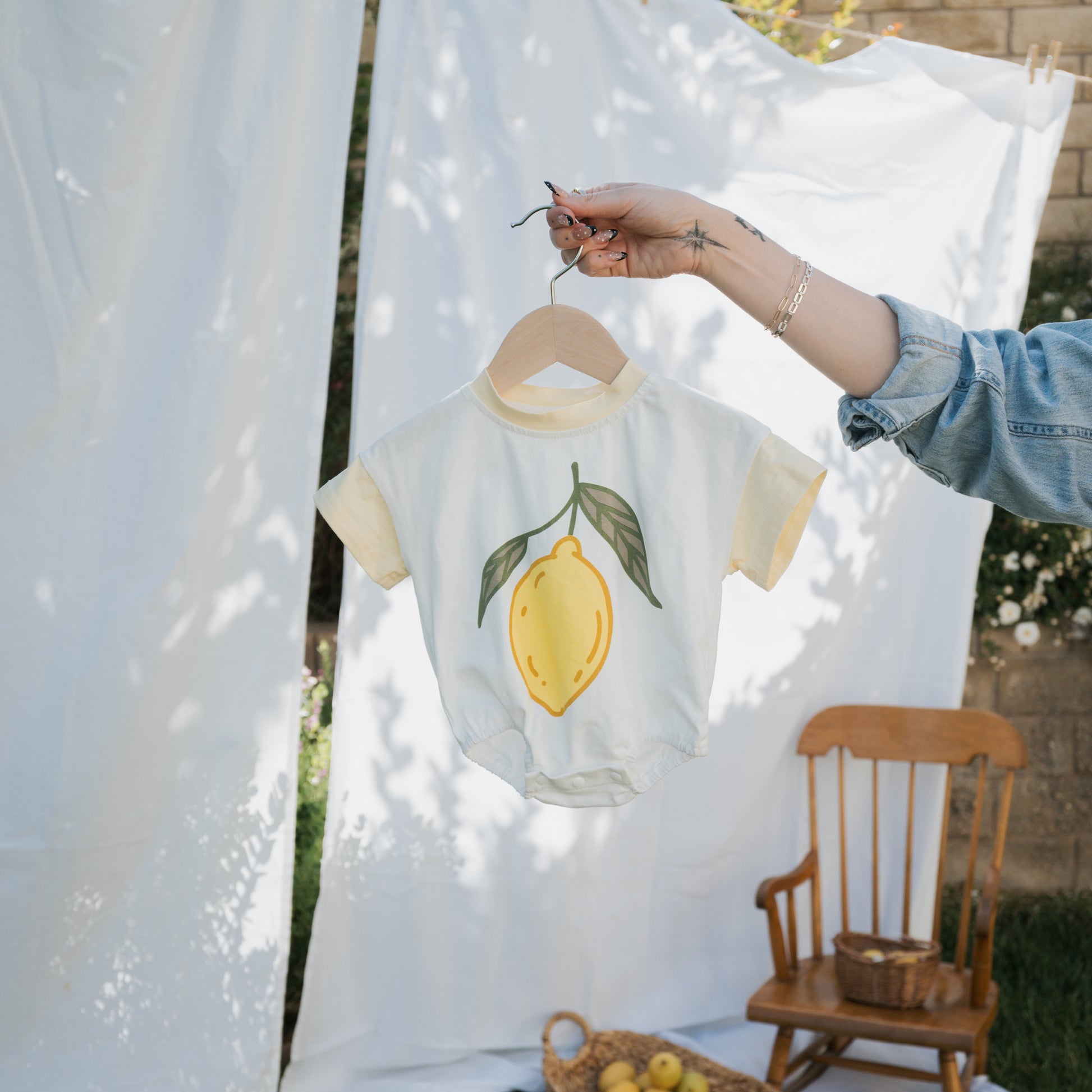 A person holds up the forever french baby Short Sleeve Romper | Lemon against an outdoor scene with white sheets, a wooden chair, and a basket of lemons—showcasing this charming summer baby outfit.