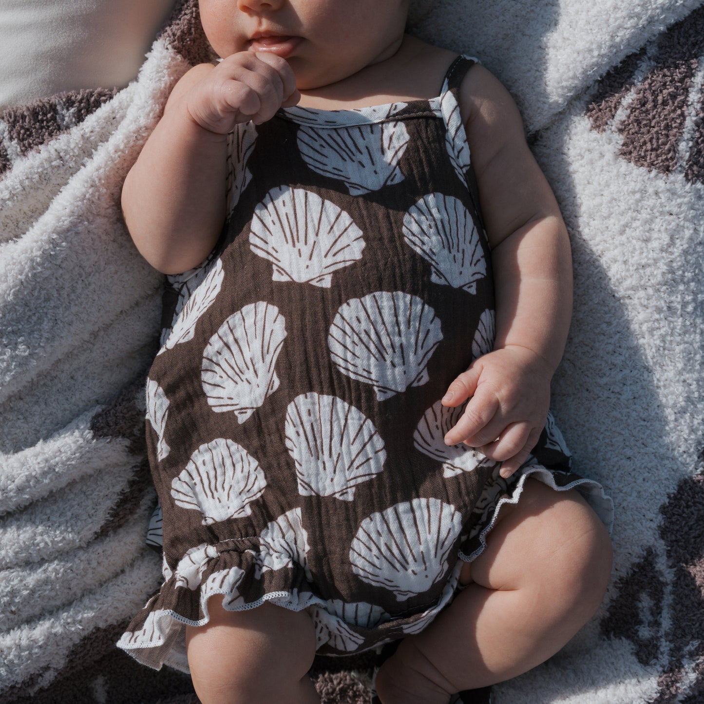 A newborn in the forever french baby Muslin Sleeveless Tank Romper | Seashells lies on a soft white and brown textured blanket, with one hand near their mouth and the other by their side.