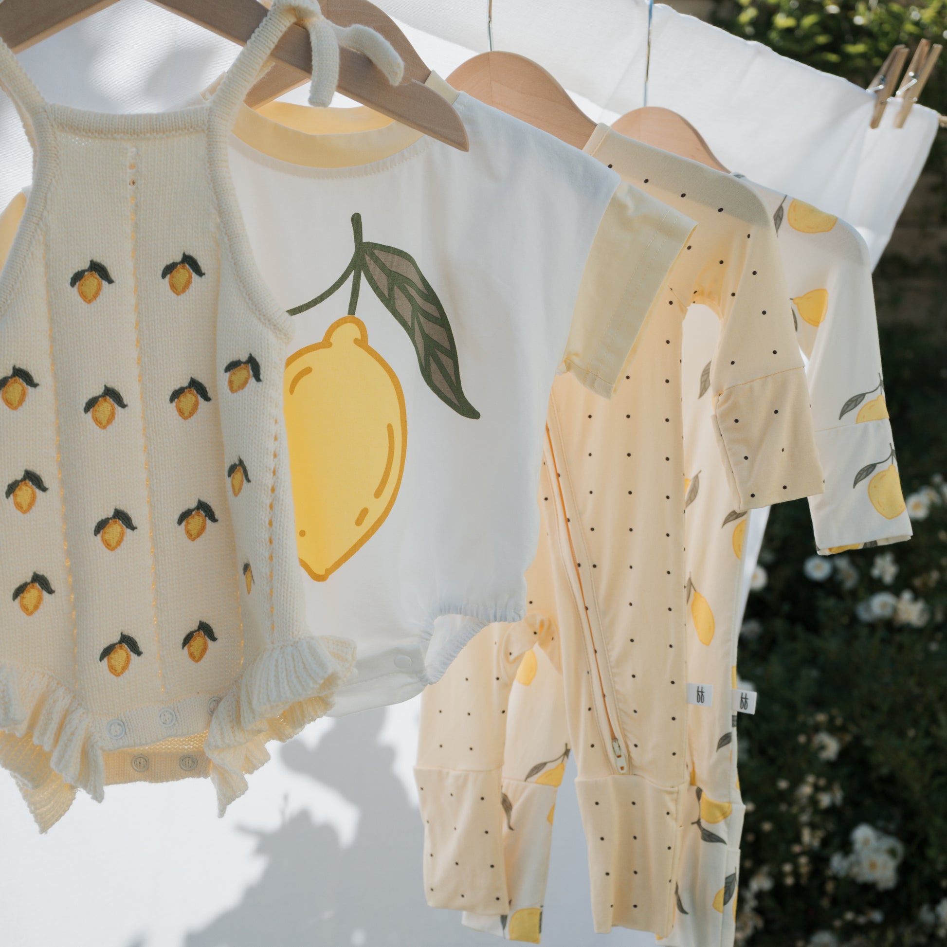 Four baby outfits, including the forever french baby Short Sleeve Romper in Lemon, hang on wooden hangers outdoors amid sunlight and greenery—perfect inspiration for a playful summer look.
