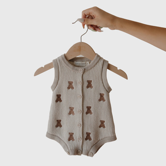 A hand holds the forever french baby Knit Romper | Teddy, a sleeveless beige 100% cotton romper with embroidered teddy bears and front buttons, displayed on a wooden hanger against a plain white background.