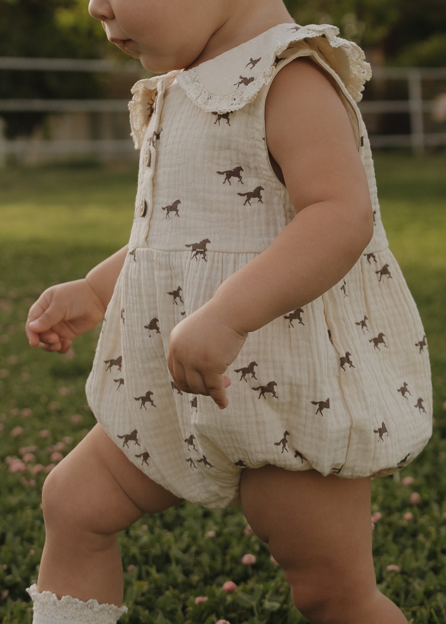 A toddler in the forever french baby Muslin Romper | Wild Horses, with only their lower face and body visible, walks on grass outdoors—showcasing this adorable piece of newborn clothing.