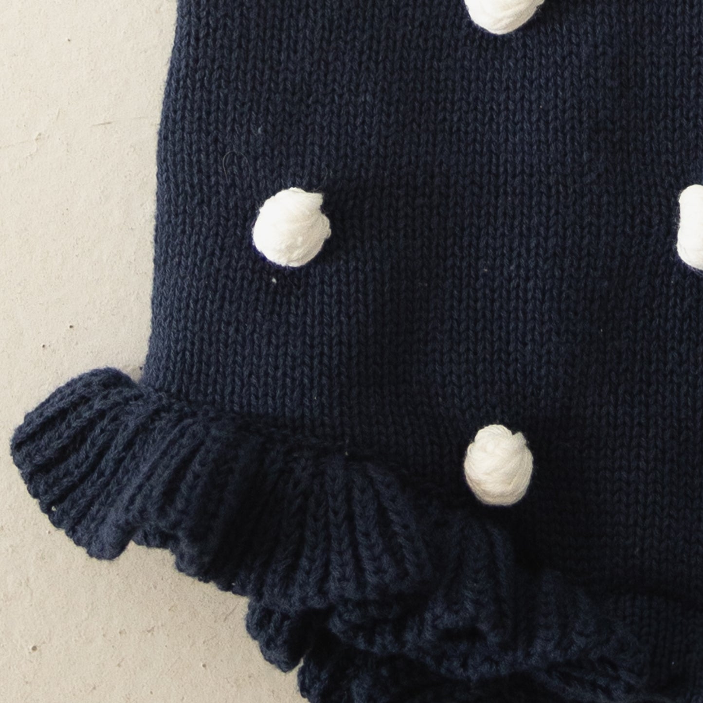 Close-up of the Knit Ruffle Romper | Emery by forever french baby, featuring dark blue soft knit with white pom-pom dots, ruffled edges, and adjustable tie straps, displayed on a light beige background.
