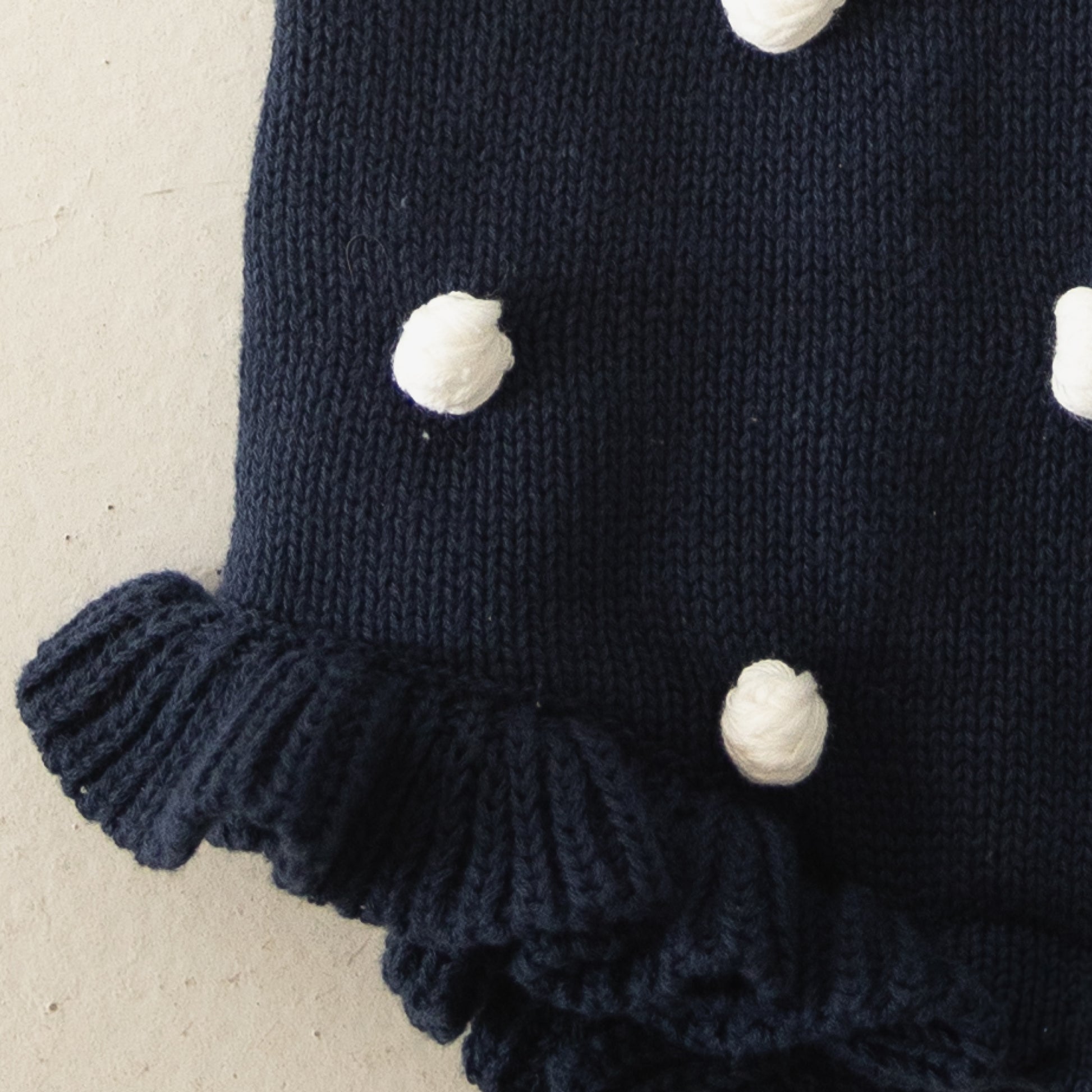 Close-up of the Knit Ruffle Romper | Emery by forever french baby, featuring dark blue soft knit with white pom-pom dots, ruffled edges, and adjustable tie straps, displayed on a light beige background.