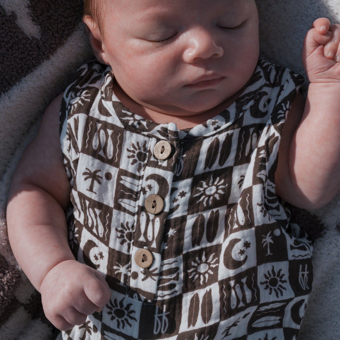 A newborn sleeps on a soft blanket wearing the forever french baby Muslin Tank Romper | Summer Check—a 100% cotton black and white check sleeveless romper with wooden buttons, one hand gently curled.