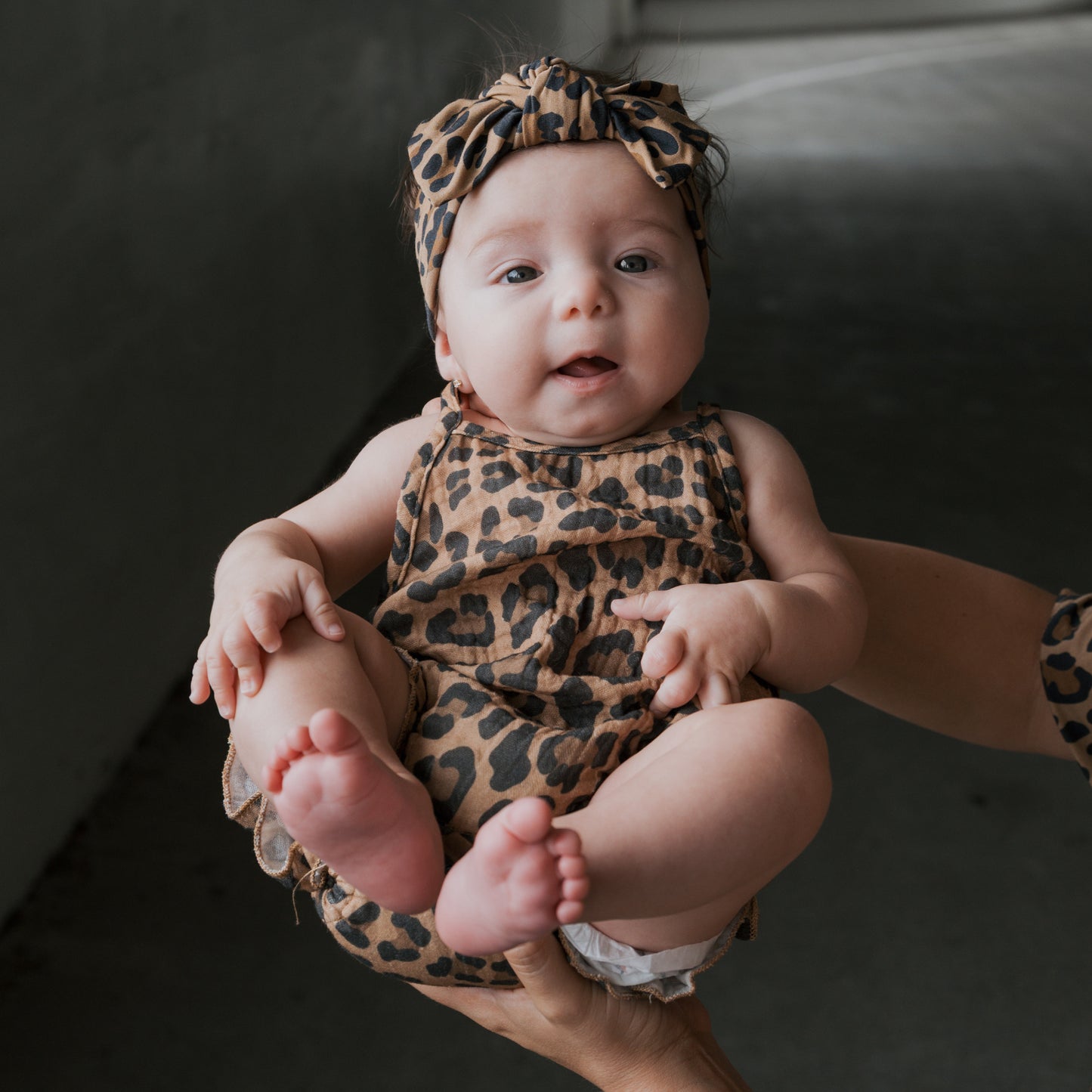 A baby wearing the Muslin Sleeveless Tank Romper in Leopard from forever french baby, with a matching headband, is held up by adult hands and gazes curiously at the camera—a delightful moment.