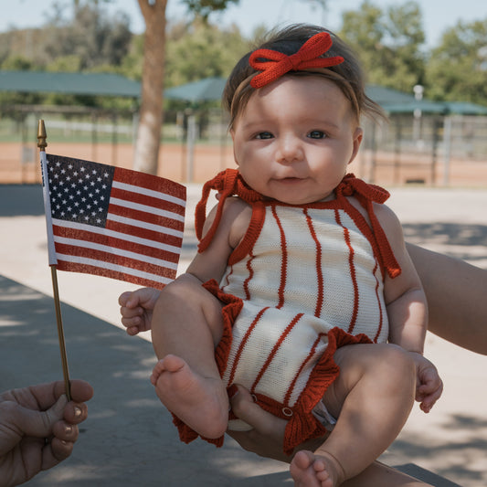 A baby in the forever french baby Knit Ruffle Romper | Picnic Pinstripe and a red headband is held outdoors while someone waves a small American flag nearby. Trees and park structures appear in the background.