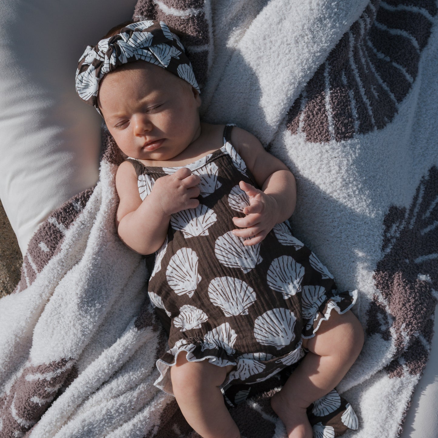 A baby sleeps peacefully on a patterned blanket, dressed in the forever french baby Muslin Sleeveless Tank Romper | Seashells, with sunlight gently illuminating the cozy scene.