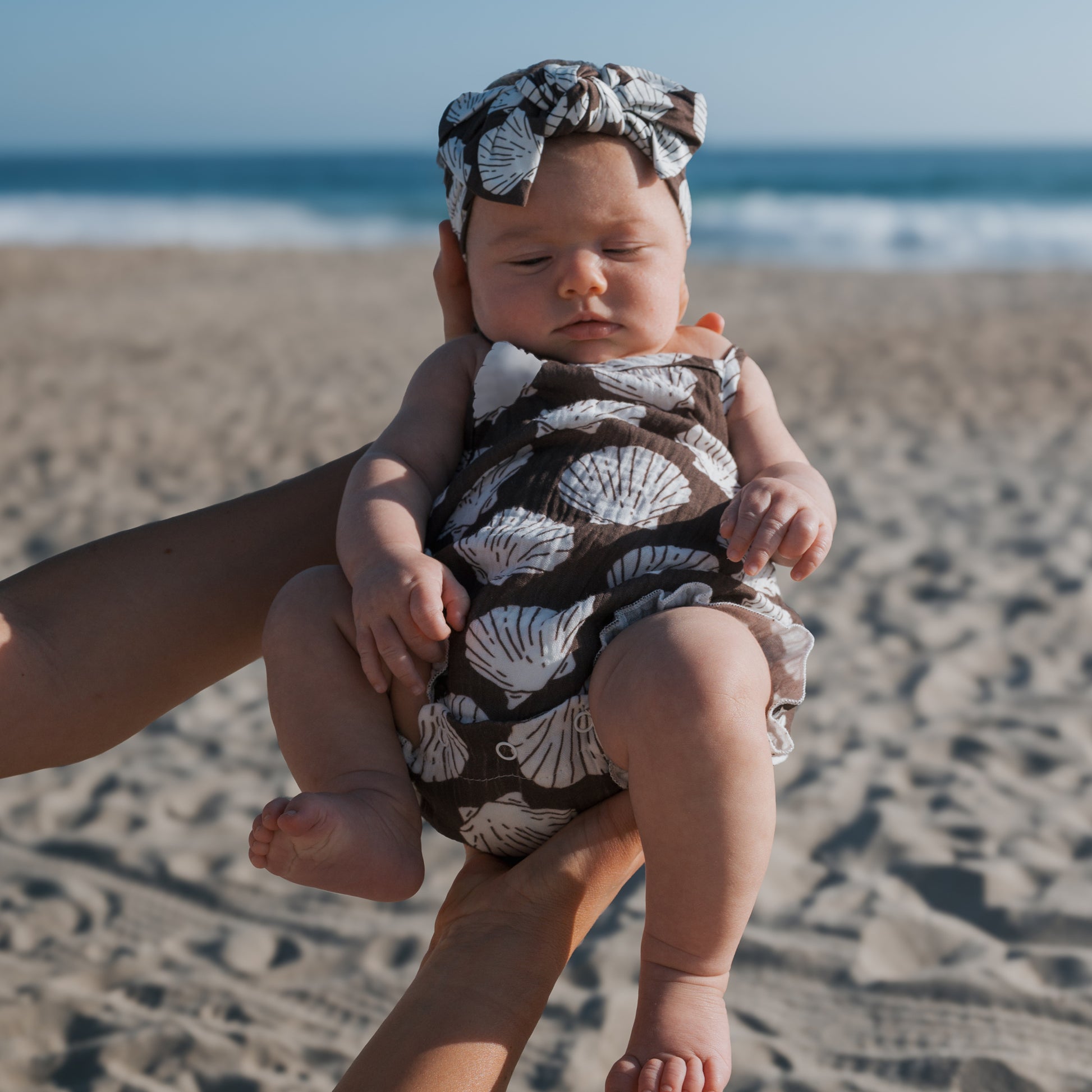 A baby in a forever french baby muslin sleeveless tank romper in seashells print is held on a sandy beach, waves and blue sky behind—perfect for coordinating family outfits.