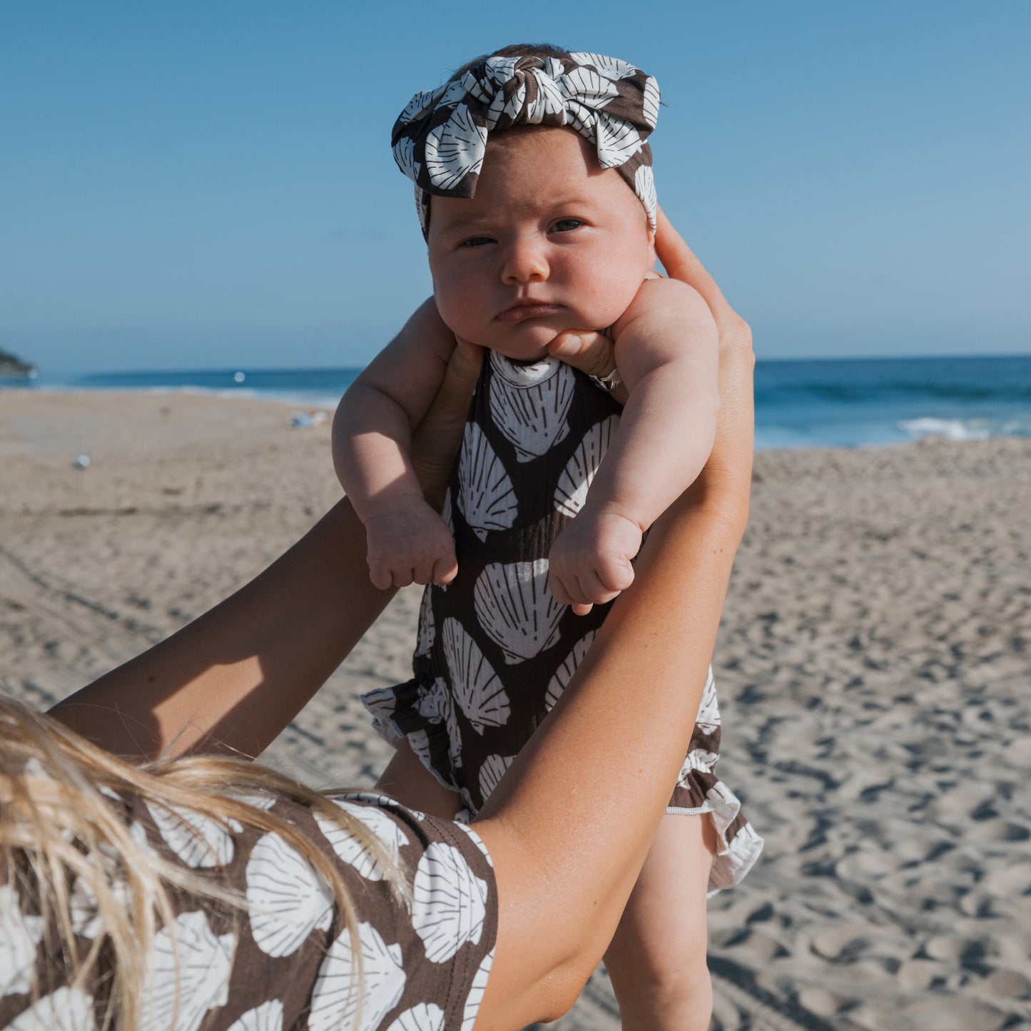 A baby in the forever french baby Muslin Sleeveless Tank Romper | Seashells is held by an adult on a sandy beach under a clear blue sky.