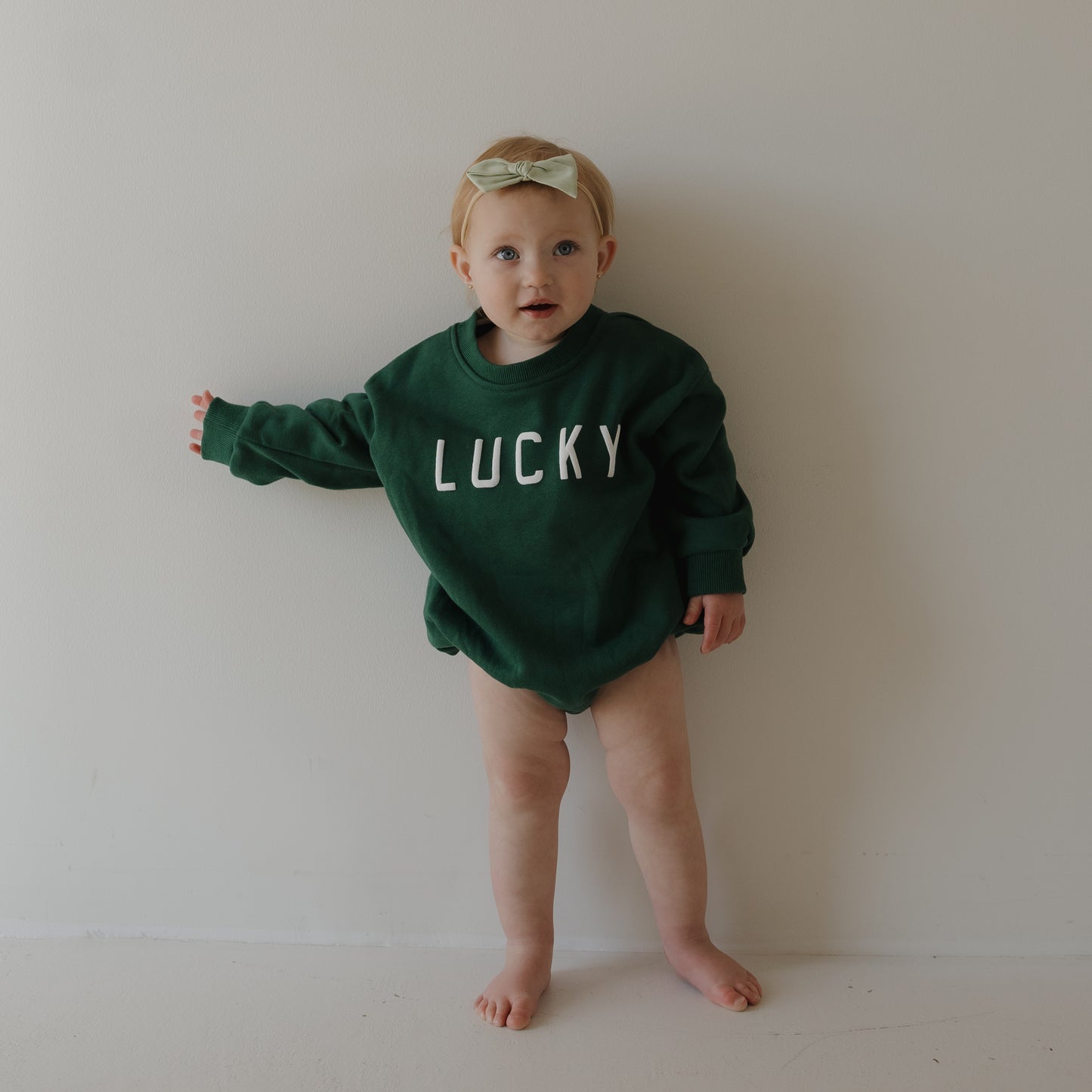 A toddler in a forever french baby "Lucky" Sweatshirt Romper—perfect for St. Patrick’s Day—and a light green bow headband stands barefoot against a plain white wall.