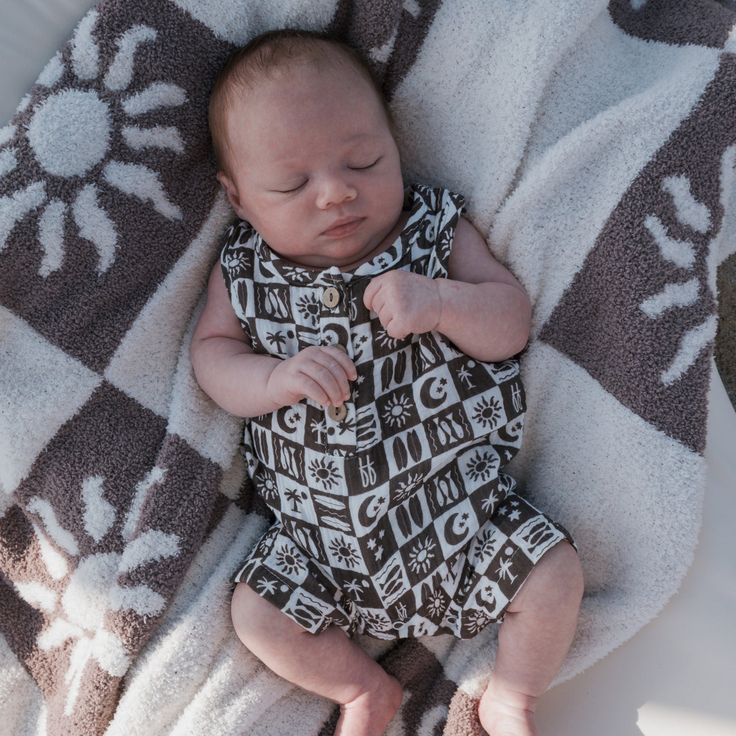 A newborn sleeps on a sun-patterned towel, wearing the forever french baby Muslin Tank Romper | Summer Check—a 100% cotton black and white summer outfit.
