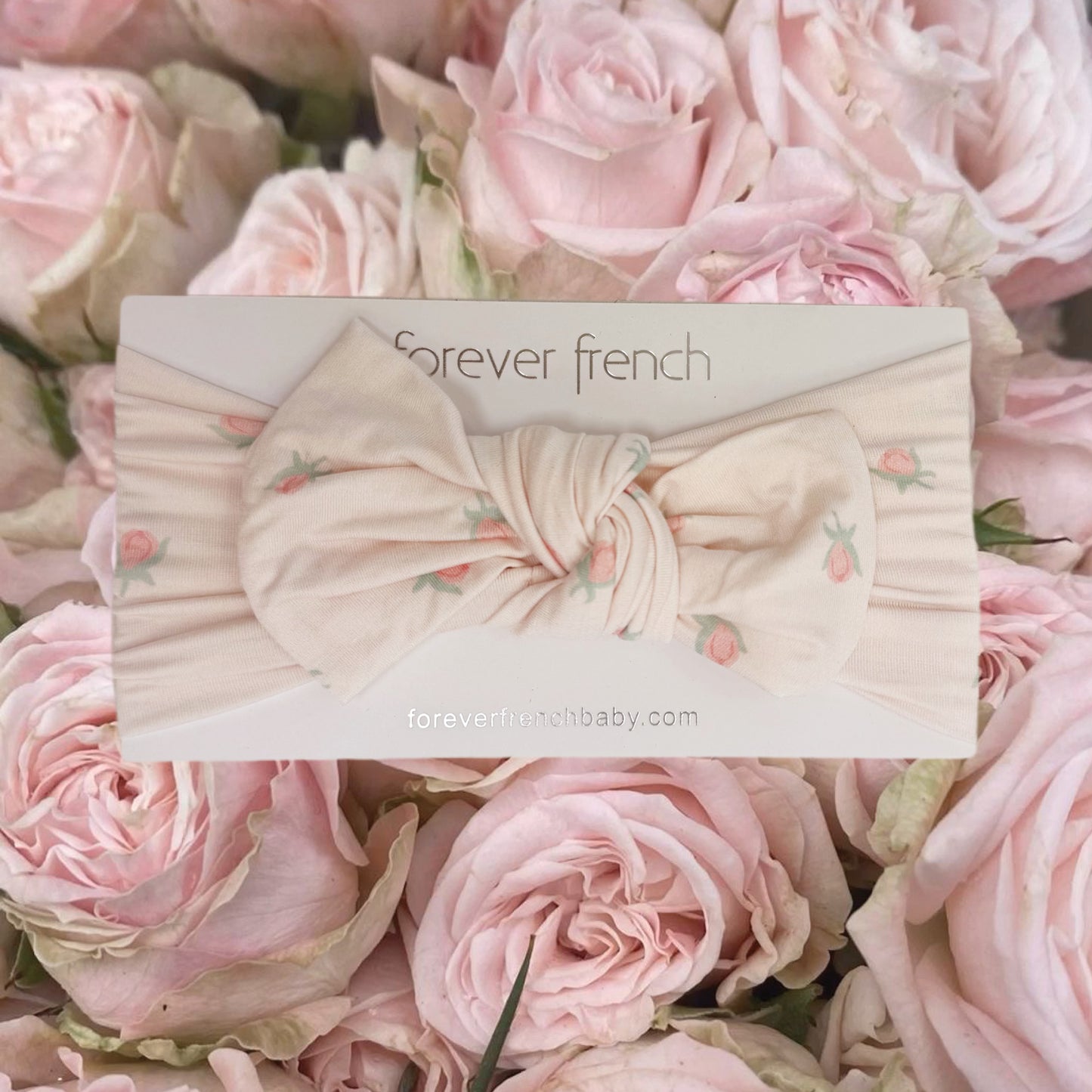 A cream-colored Bamboo Head Wrap | Rosie from forever french baby, featuring small pink floral prints, is displayed on a “forever french” card and surrounded by light pink roses. This sweet, adjustable head wrap is perfect for delicate little heads and adds a charming touch to any outfit.