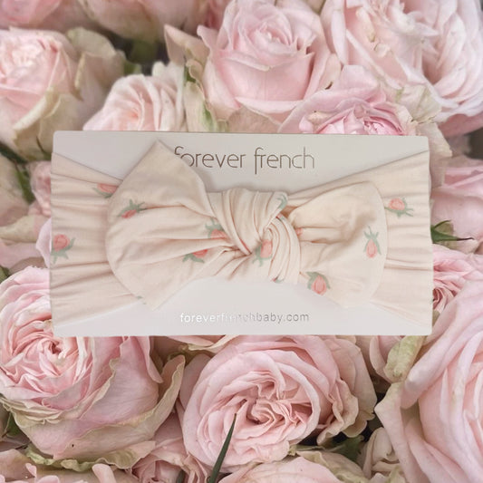 A cream-colored Bamboo Head Wrap | Rosie from forever french baby, featuring small pink floral prints, is displayed on a “forever french” card and surrounded by light pink roses. This sweet, adjustable head wrap is perfect for delicate little heads and adds a charming touch to any outfit.