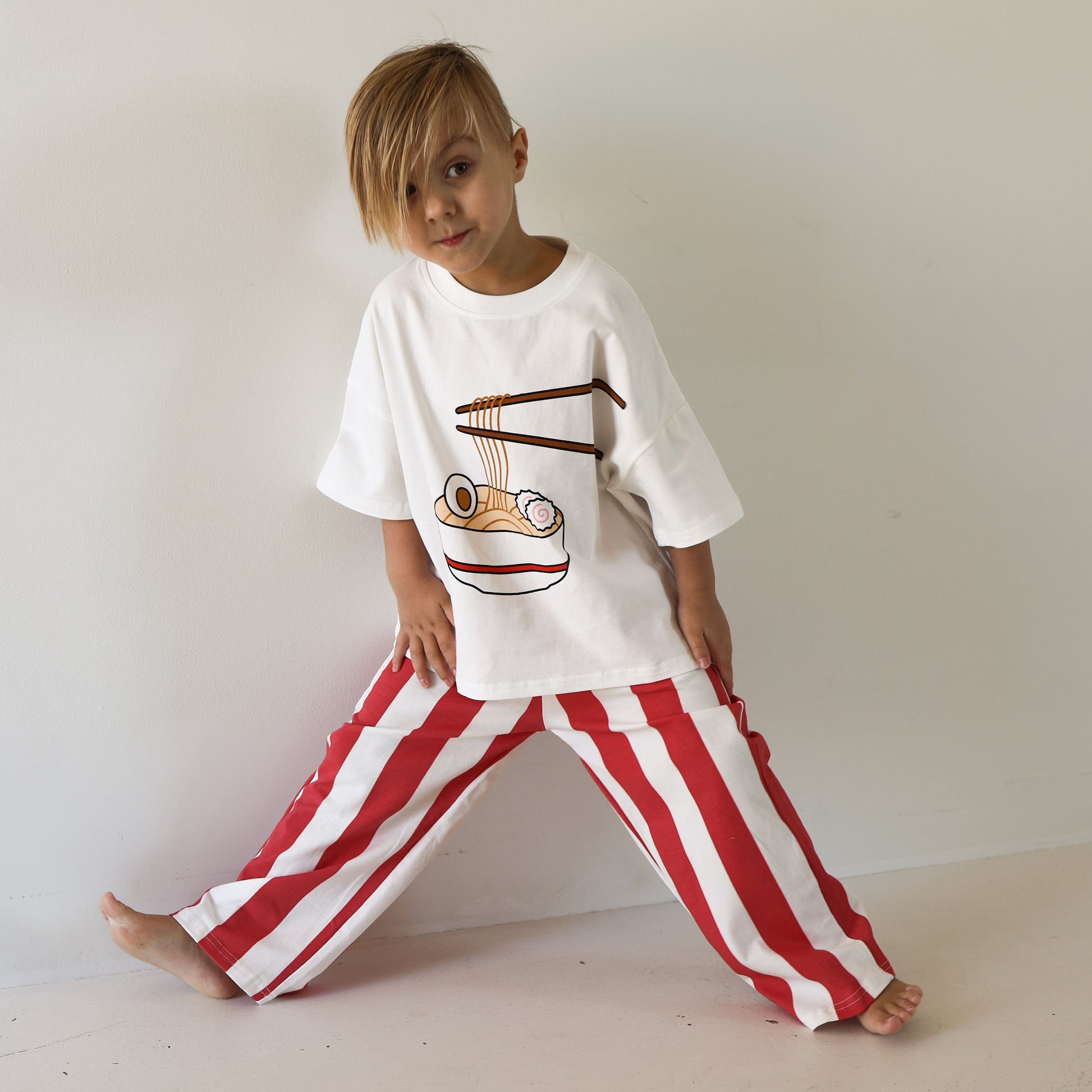 A young child with side-swept blond hair poses against a white wall, wearing the forever french baby T-Shirt & Pant Set | Ramen, featuring a cotton tee with a ramen bowl graphic and red-and-white striped pants.