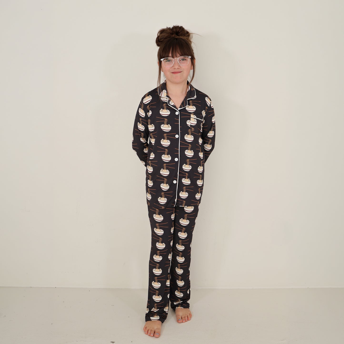 A young girl with glasses and long brown hair in a bun stands barefoot against a plain wall, smiling slightly. She wears the forever french baby Girl's Bamboo Button Up Lounge Set | Ramen, with her hands behind her back.