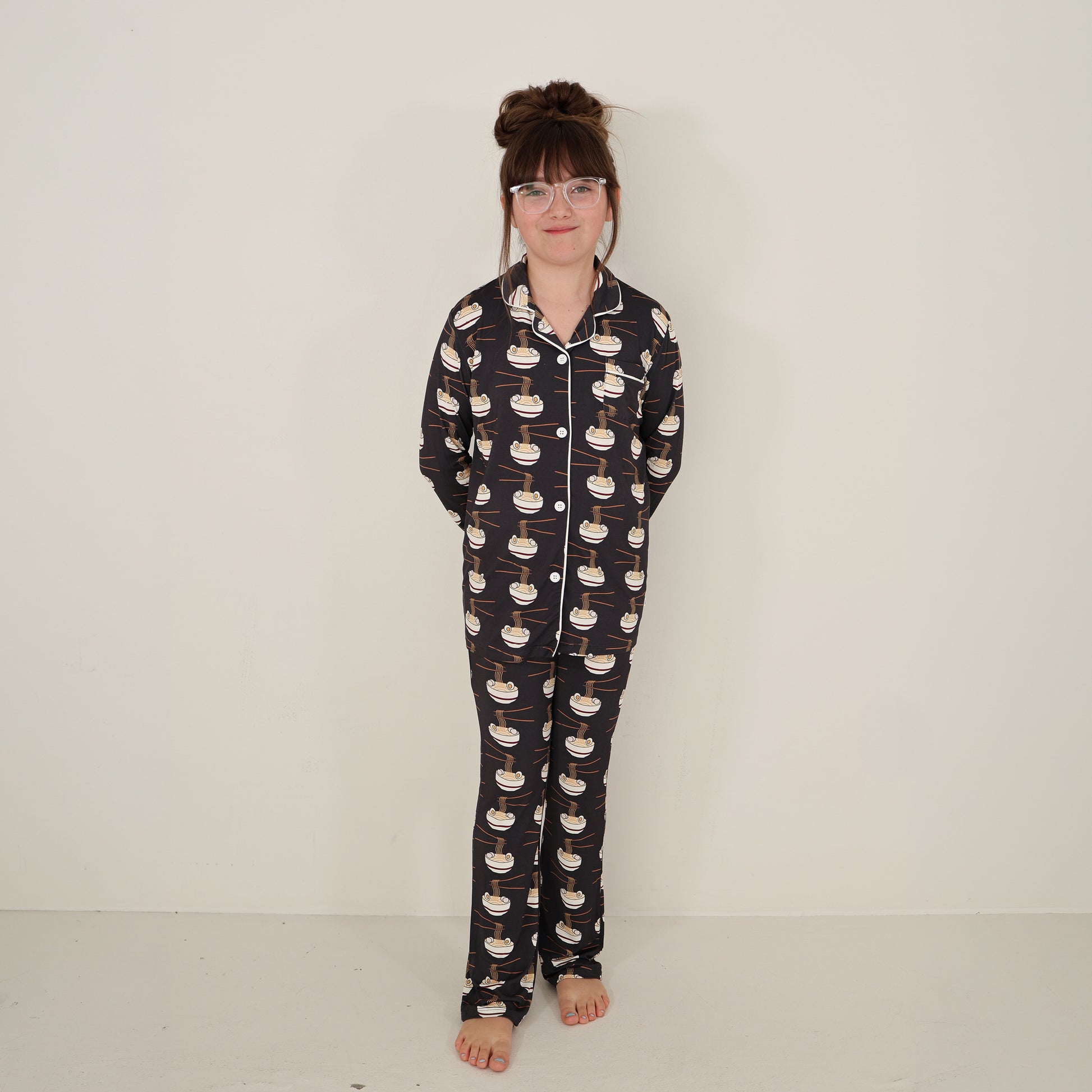 A young girl with glasses and long brown hair in a bun stands barefoot against a plain wall, smiling slightly. She wears the forever french baby Girl's Bamboo Button Up Lounge Set | Ramen, with her hands behind her back.