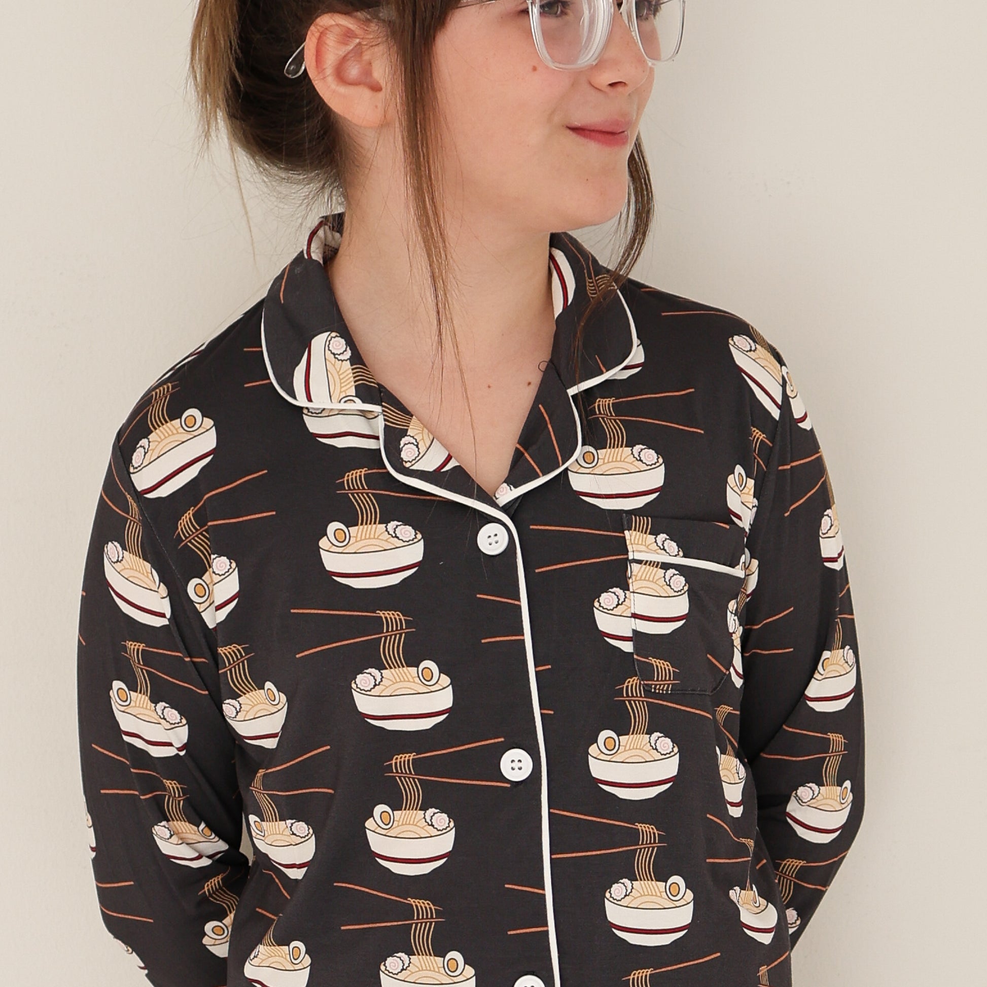 A person wearing glasses and the forever french baby Girl's Bamboo Button Up Lounge Set | Ramen, featuring a ramen bowl and chopsticks pattern, stands against a plain light-colored wall, looking to the side with a slight smile.