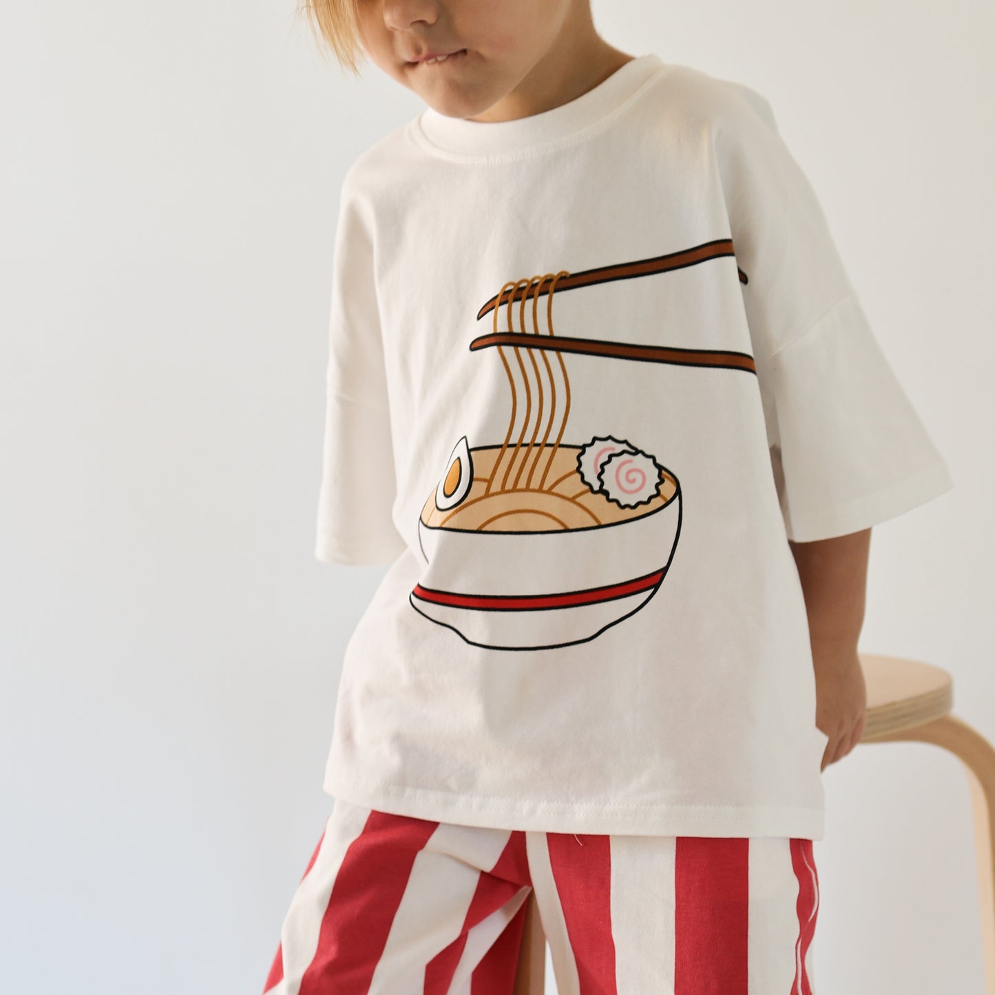 A child models the forever french baby T-Shirt & Pant Set | Ramen, featuring a cotton tee with a cartoon ramen bowl and chopsticks graphic, paired with red and white striped pants—a playful kids’ daywear set.