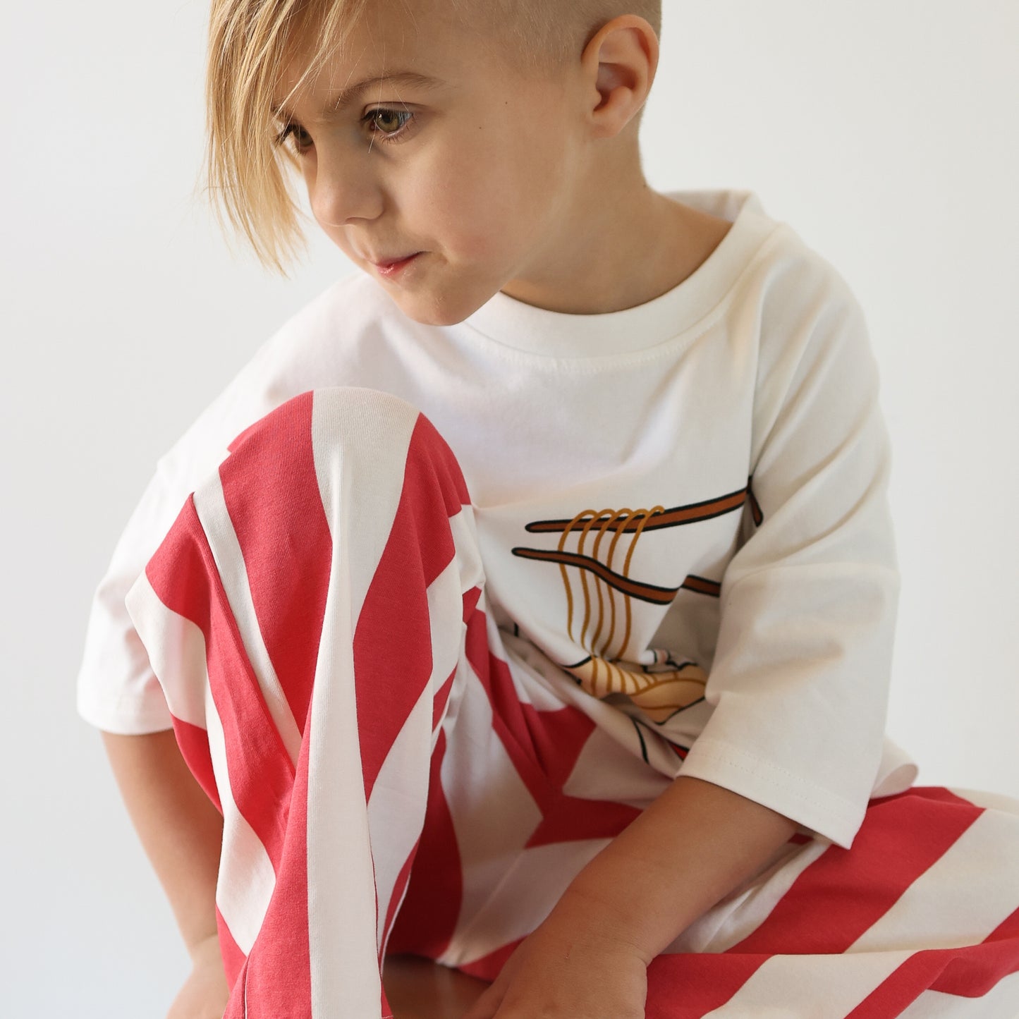A young child with blonde hair and a side shave sits with one knee up, wearing the forever french baby T-Shirt & Pant Set | Ramen: a white graphic tee and red-and-white striped pants, looking off to the side against a plain background.