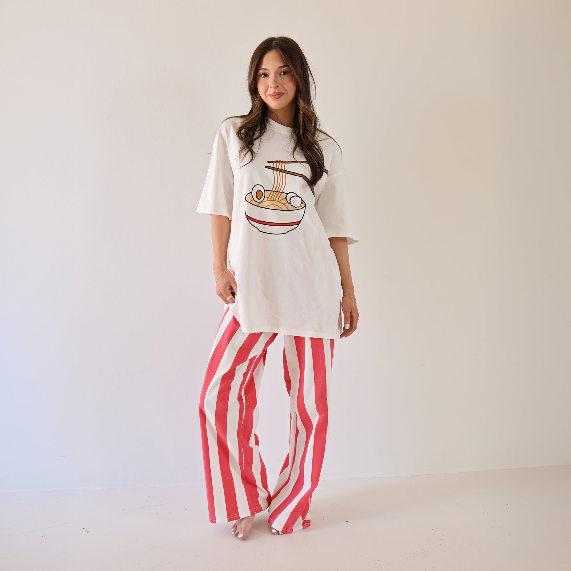 A woman smiles barefoot against a white wall, wearing a white tee with a burger graphic and forever french baby Women's Wide Leg Pants | Tokyo Stripe—red-and-white vertical stripes and an elastic waistband. Her long brown hair flows freely.