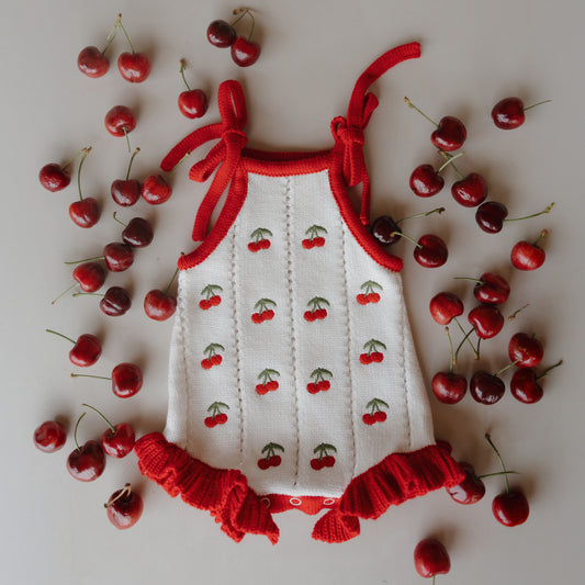 The Knit Ruffle Romper | Cherry by forever french baby features charming cherry patterns, adjustable tie straps, and red ruffled edges. The cotton romper is styled with fresh cherries scattered on a light background.