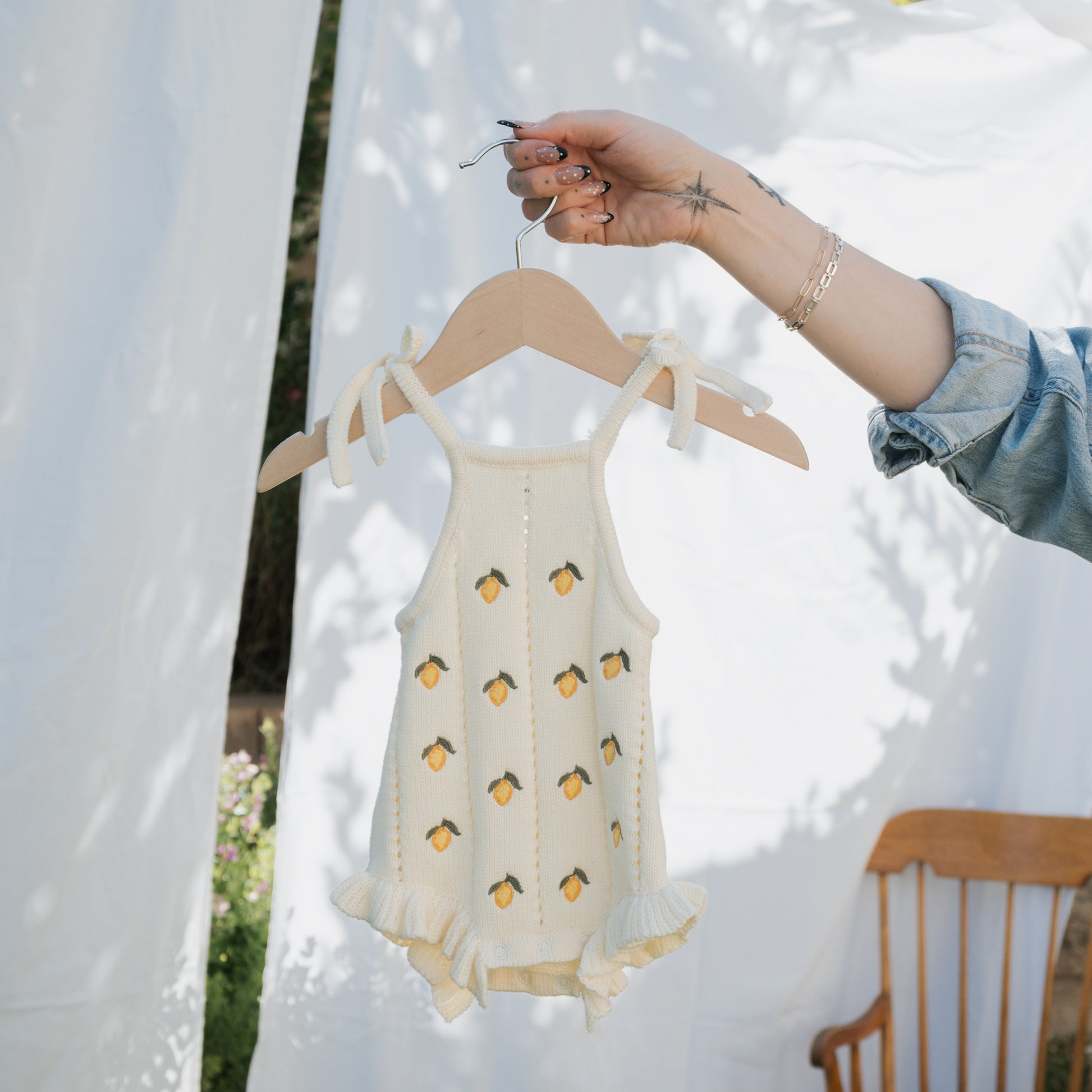 A person with tattooed fingers holds up the forever french baby Knit Ruffle Romper | Lemon, a cream knit romper with yellow lemon patterns, ruffled leg openings, and tie straps. A wooden chair and white fabric are in the background.