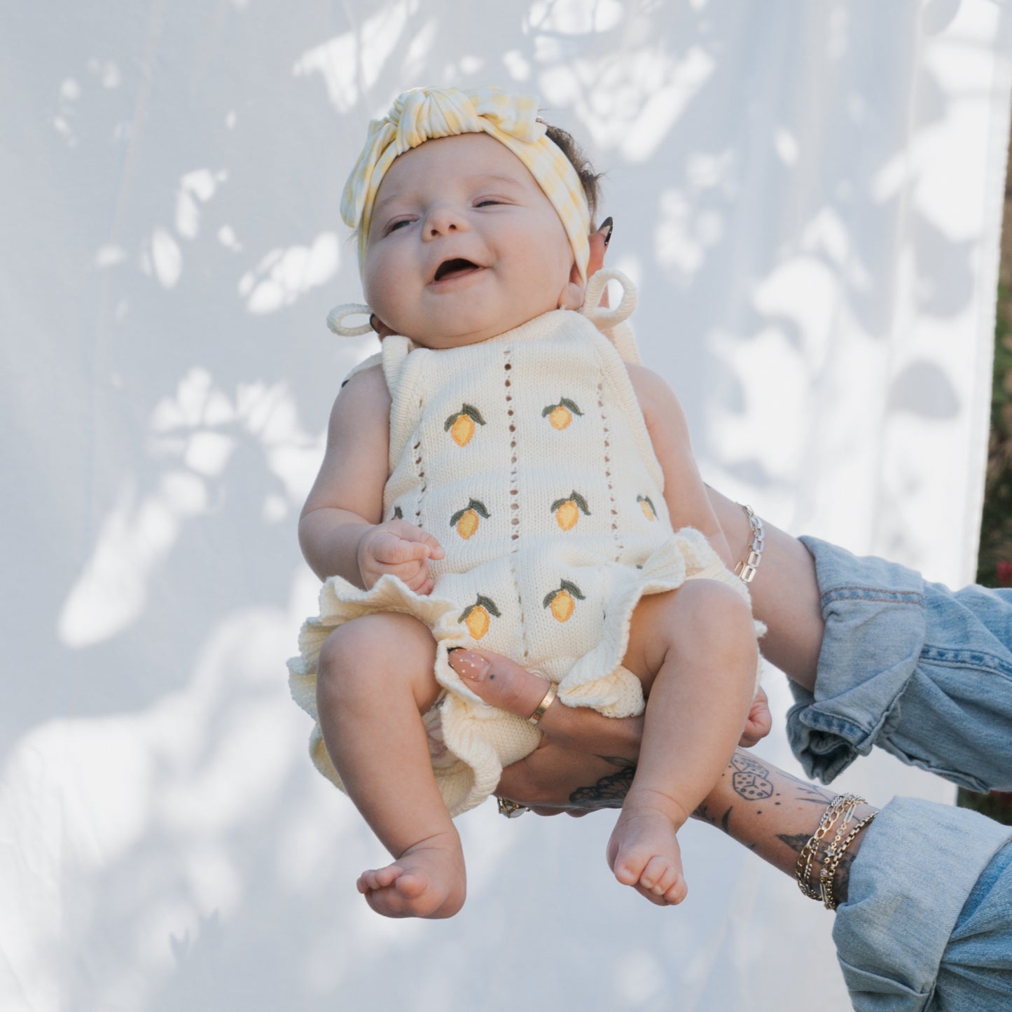 A smiling baby wears the forever french baby Knit Ruffle Romper | Lemon with adjustable tie straps and snaps, plus a matching headband, held outdoors by a tattooed adult against a sun-dappled white fabric backdrop.